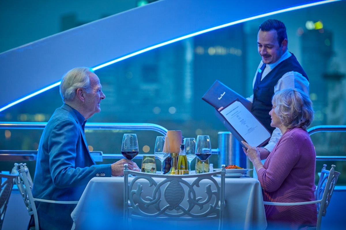 A man and a woman having dinner at a restaurant, with a waiter taking their order, in an elegant setting with a city view.