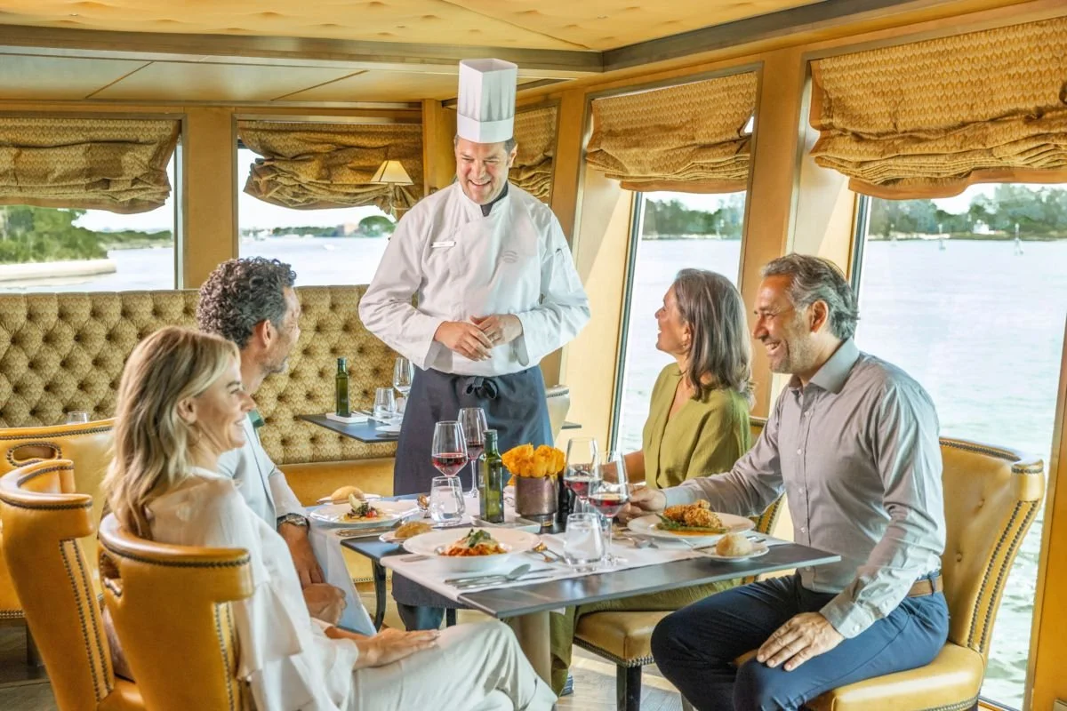 A chef standing and smiling at a group of four diners seated in a well-lit restaurant near the water, with large windows showing a lake or river view. The diners are enjoying their meal and drinks, with plates of food and glasses of wine on the table.