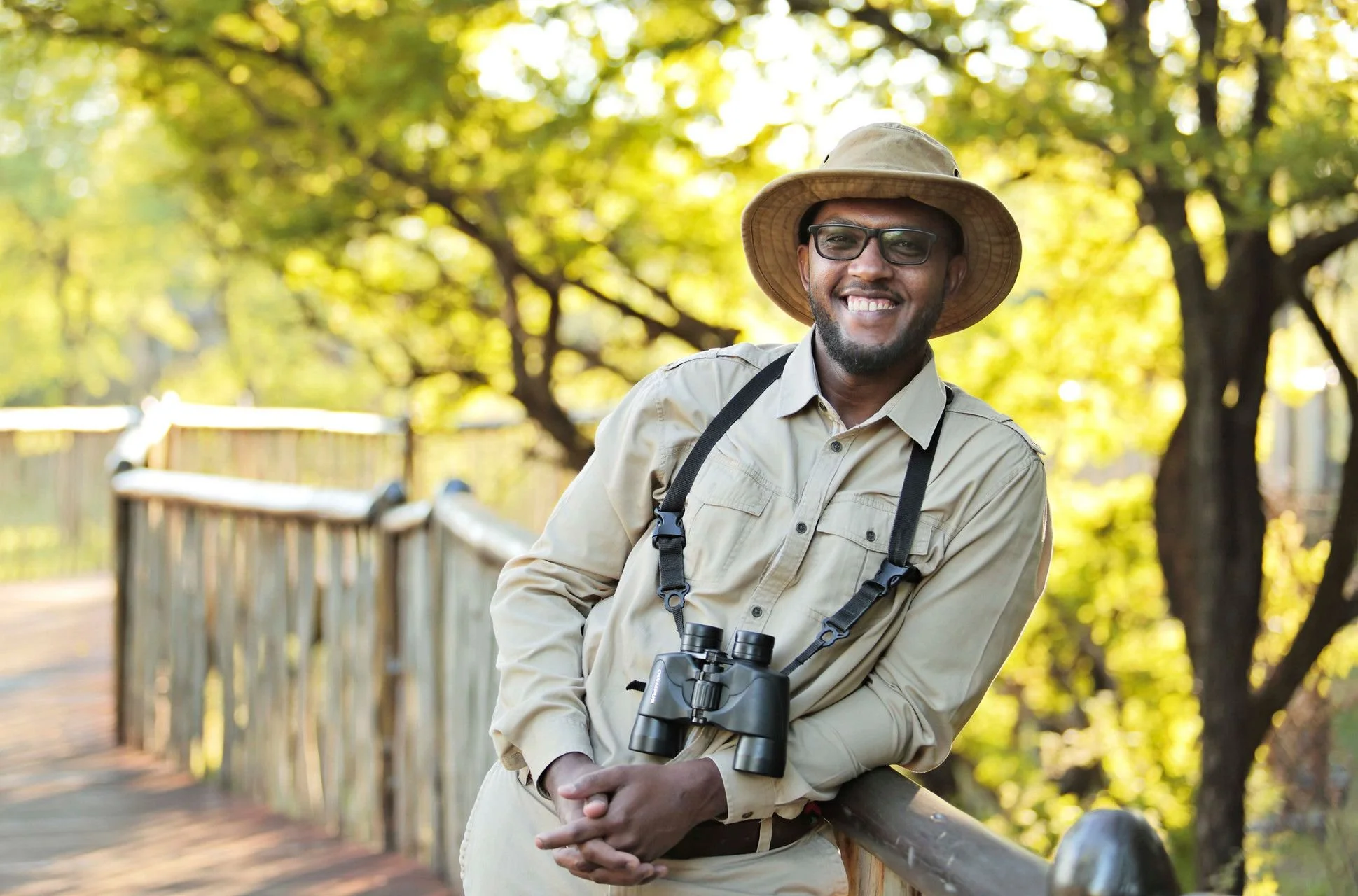 A man with glasses and a wide-brimmed hat smiling outdoors, holding binoculars, standing on a wooden bridge surrounded by trees with yellow-green leaves.