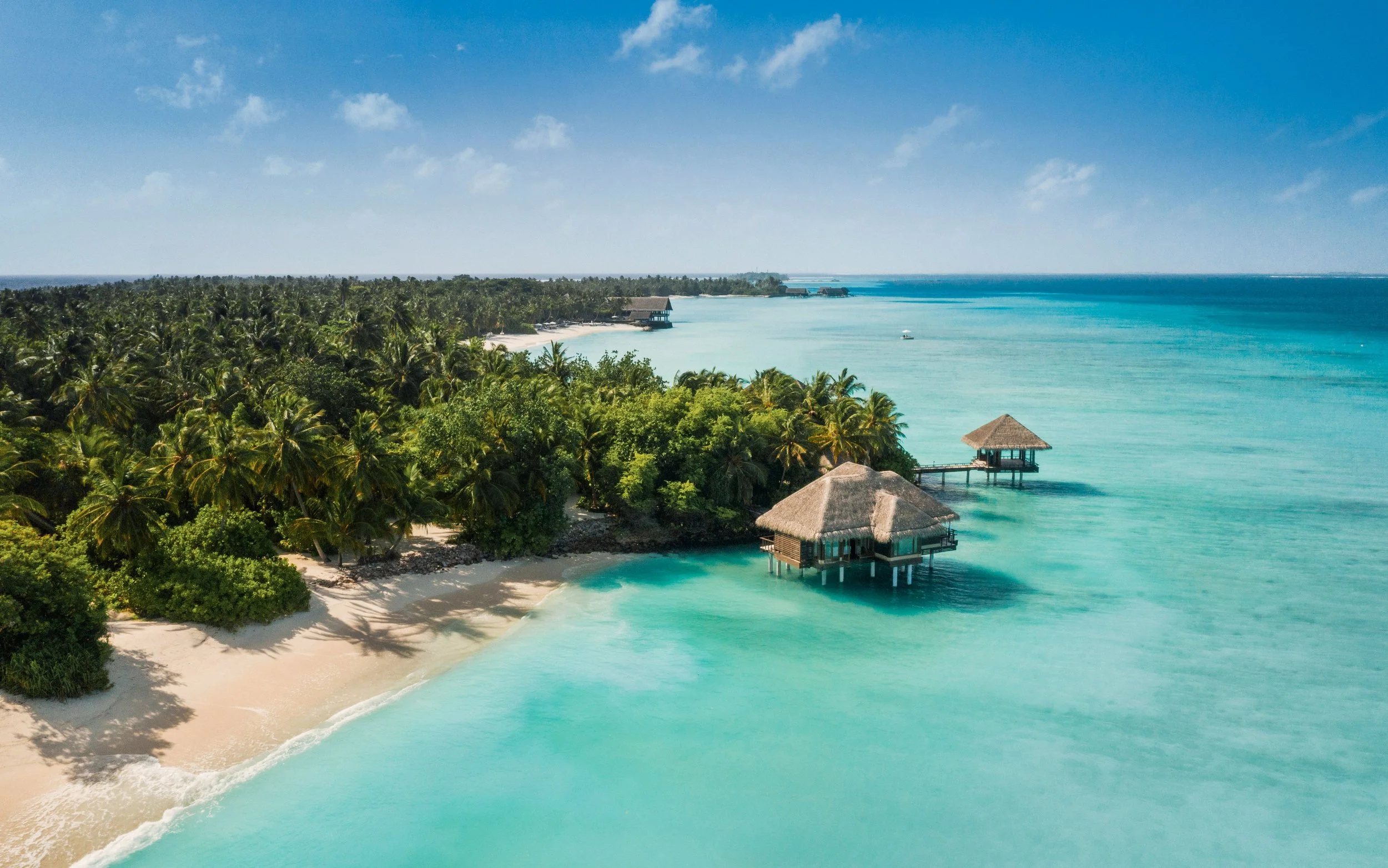 A tropical beach with white sand, clear turquoise water, lush green palm trees, and overwater bavilions, under a partly cloudy sky.
