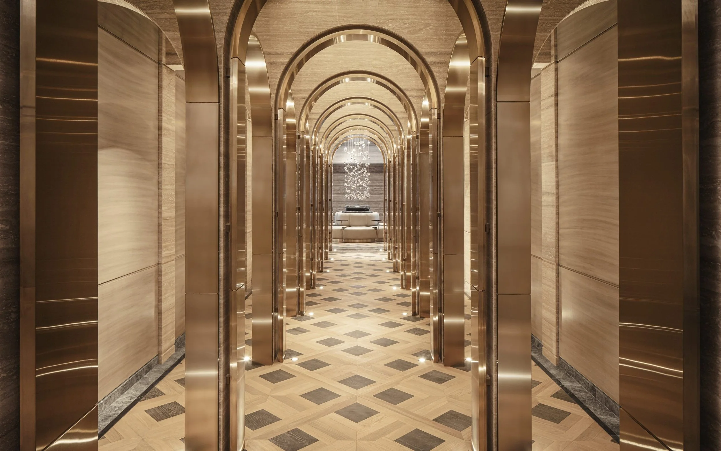 Elegant hotel corridor with golden arched doorway frames and a chandelier at the end. Marble walls and patterned wood floor.