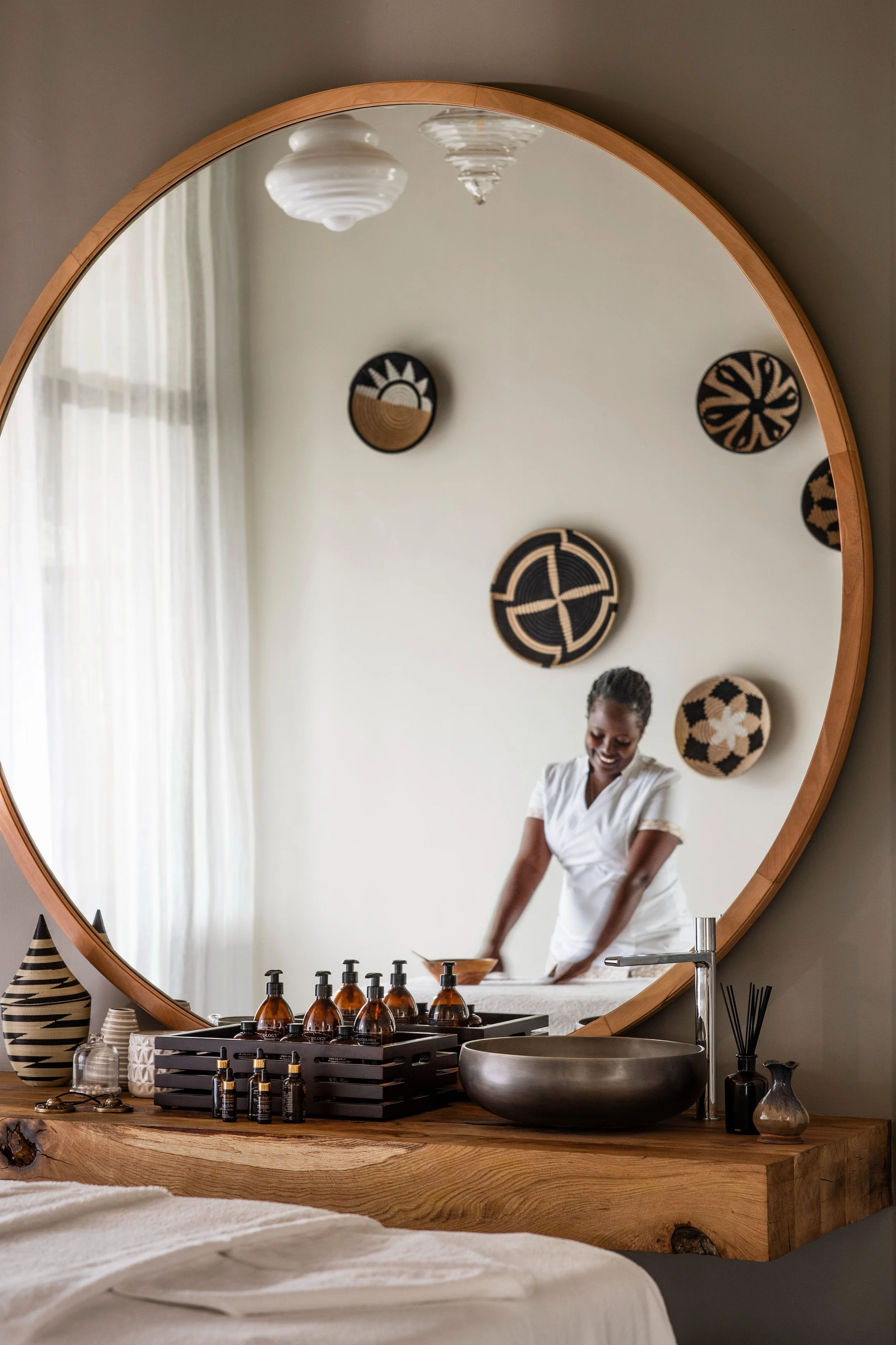 Round wall mirror reflecting a woman in a white shirt, a white curtain, wall art, and a bathroom countertop with black and amber bottles, a black tray, a black bowl, and decorative objects.