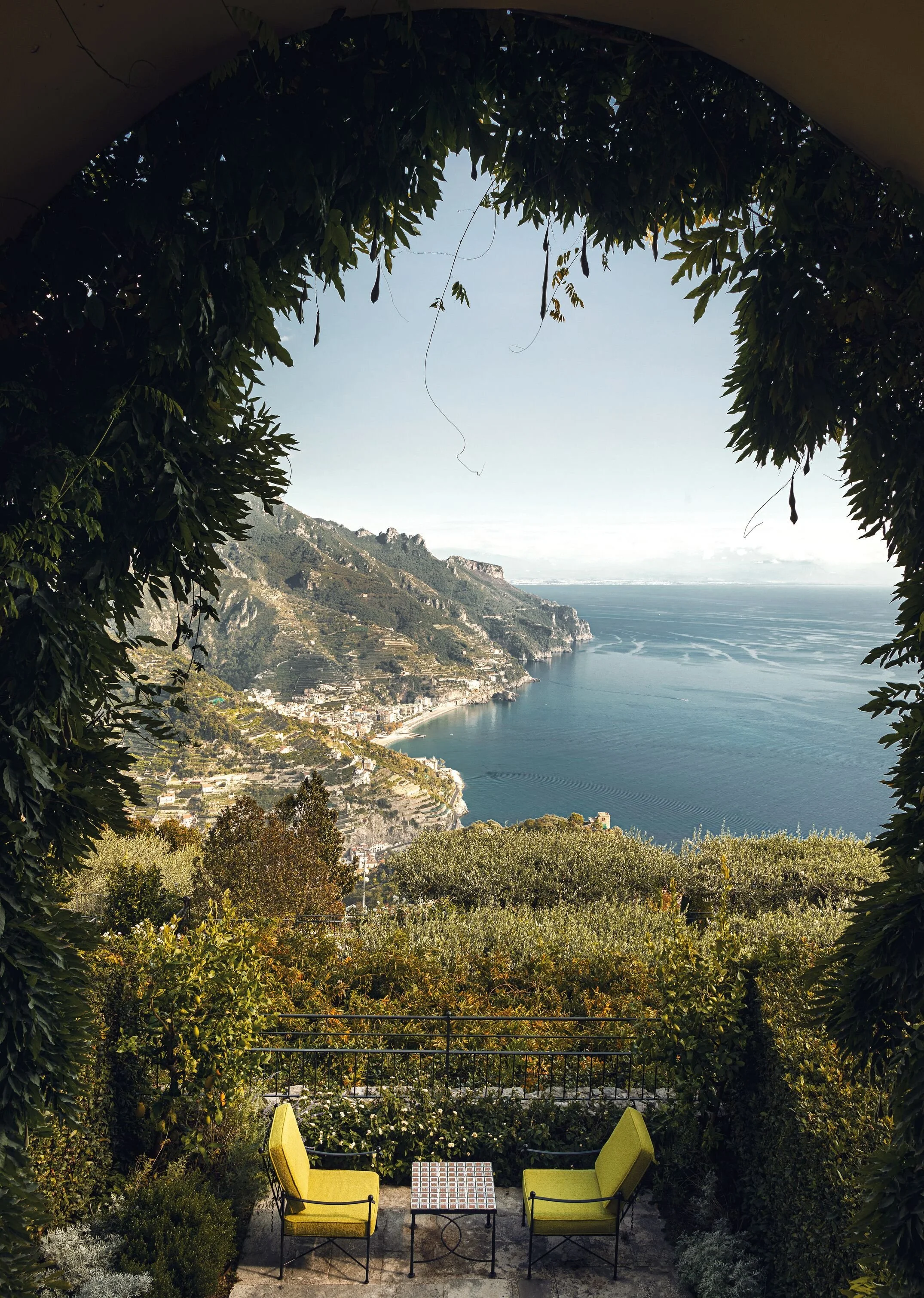 An outdoor terrace with two green chairs and a small table, surrounded by lush green plants and trees, overlooking a coastal town and mountains along the ocean under a clear sky.
