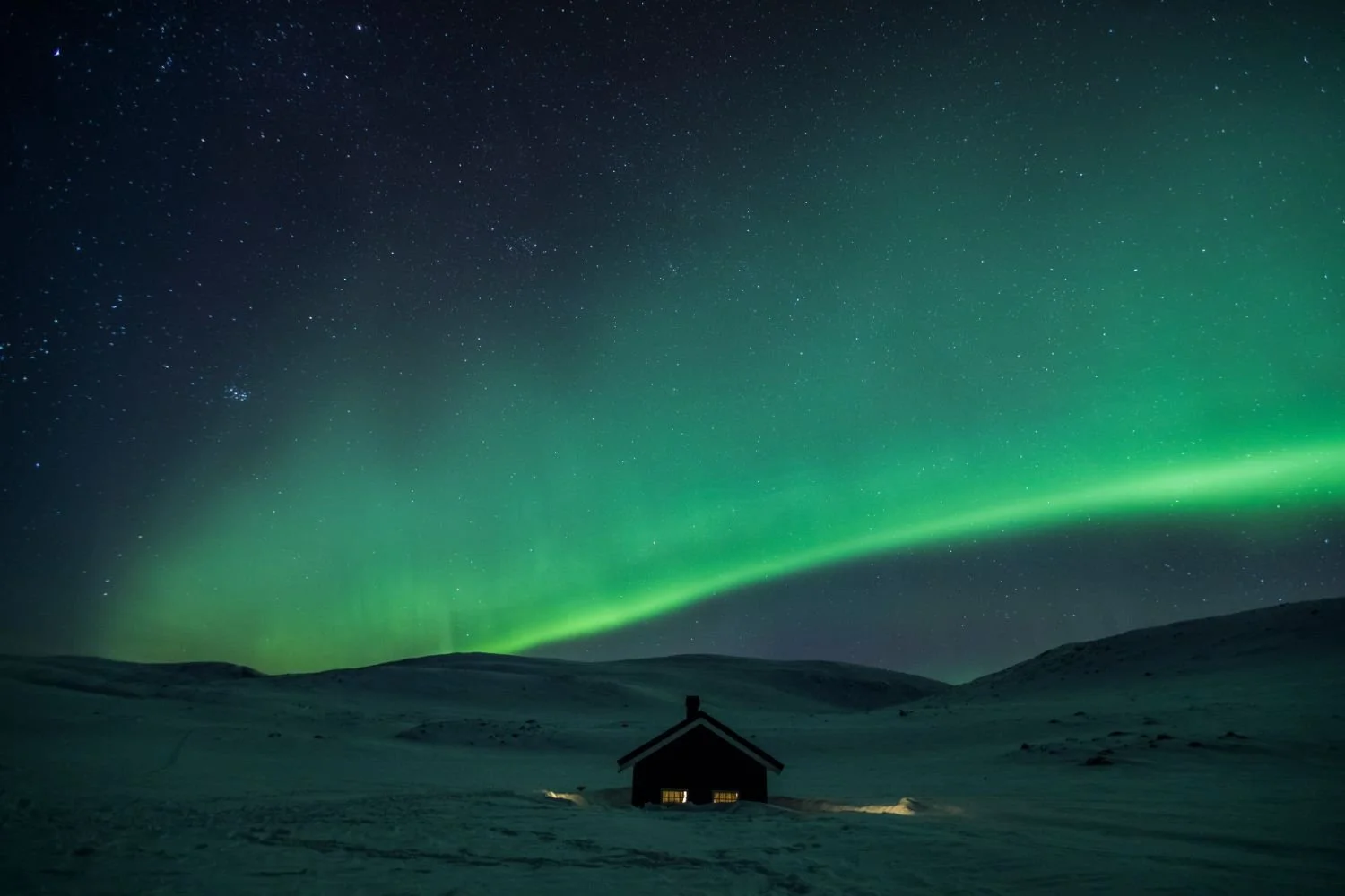 Nighttime scene with the northern lights (aurora borealis) over a snow-covered landscape and a small house with illuminated windows.
