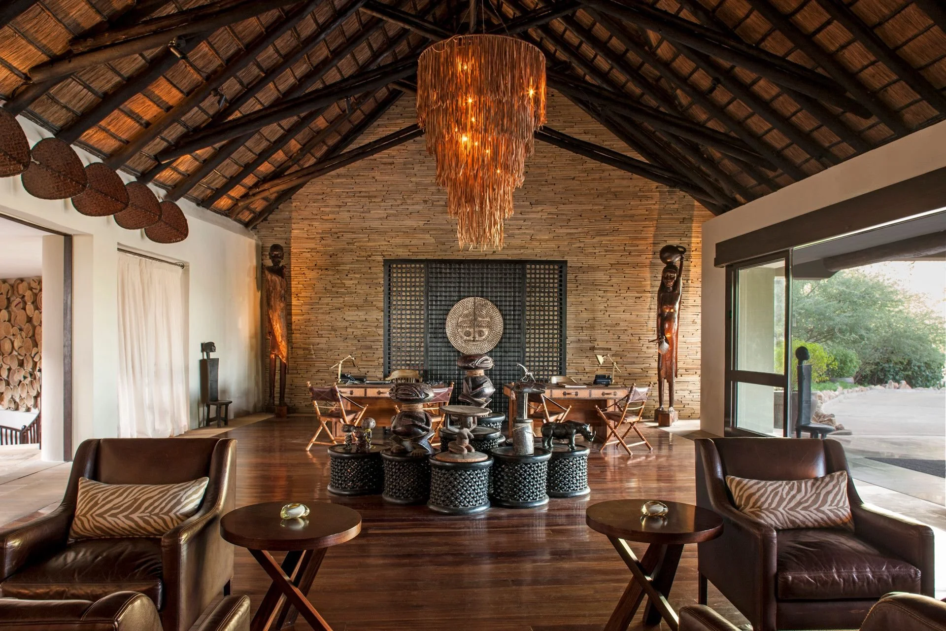 Interior of a room with a high thatched ceiling, a large hanging chandelier, wooden sculptures, and furniture including leather chairs and small tables. Large windows and a sliding door reveal an outdoor garden area.