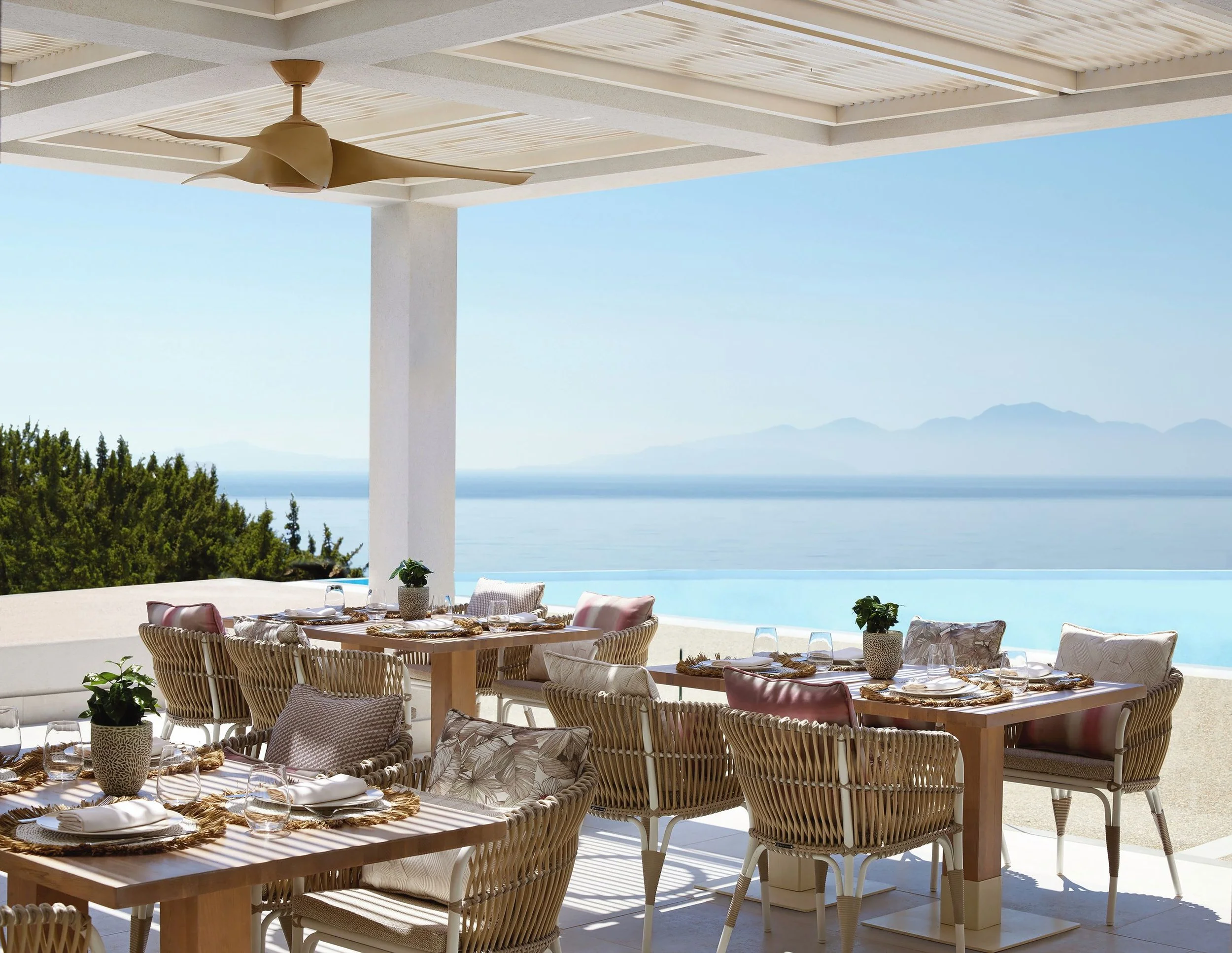 Outdoor dining area with wicker chairs and a wooden table set with plates, glasses, and small potted plants, overlooking a body of water and distant mountains under a shaded patio with a ceiling fan.