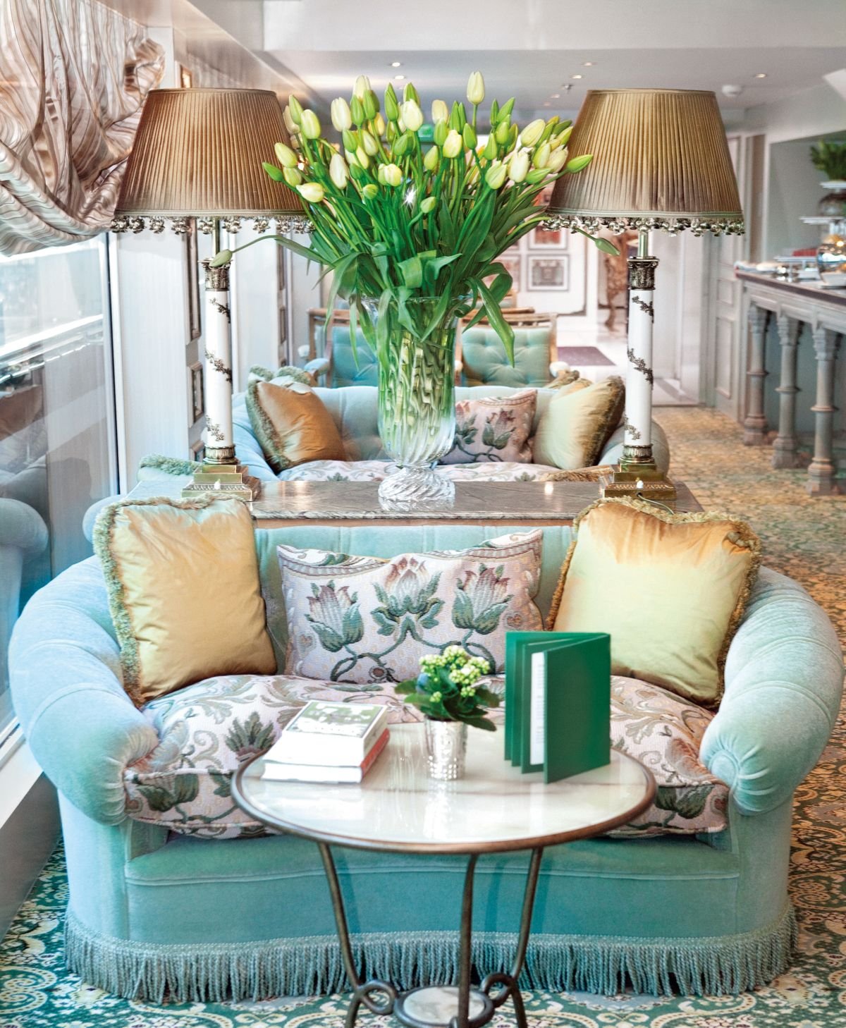 Living room with light blue upholstered sofa and circular coffee table with books and a small plant, centered around a large glass vase filled with white tulips, flanked by two elegant table lamps.