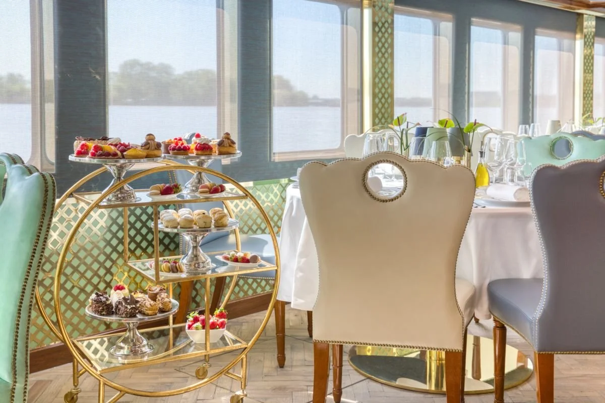 Elegant dining room with a table set for a meal, decorated with flowers and glassware, with a dessert stand displaying an assortment of pastries and desserts near large windows showing a waterfront view.