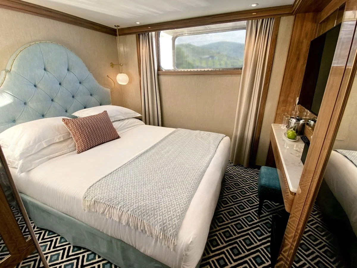 A hotel room with a large bed with a blue tufted headboard, white bedding, a patterned pillow, and a beige throw blanket. There is a window with beige curtains, a wall-mounted lamp, a marble-topped dresser, a chair, and a wall-mounted TV.