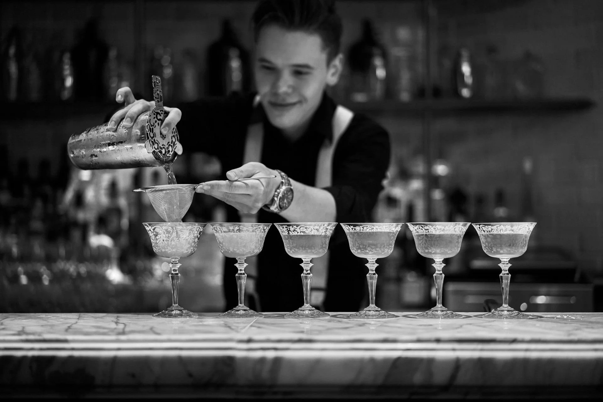 A bartender in a dark shirt and apron is pouring liquid into ornate cocktail glasses arranged in a row on a bar counter, with bottles in the background.