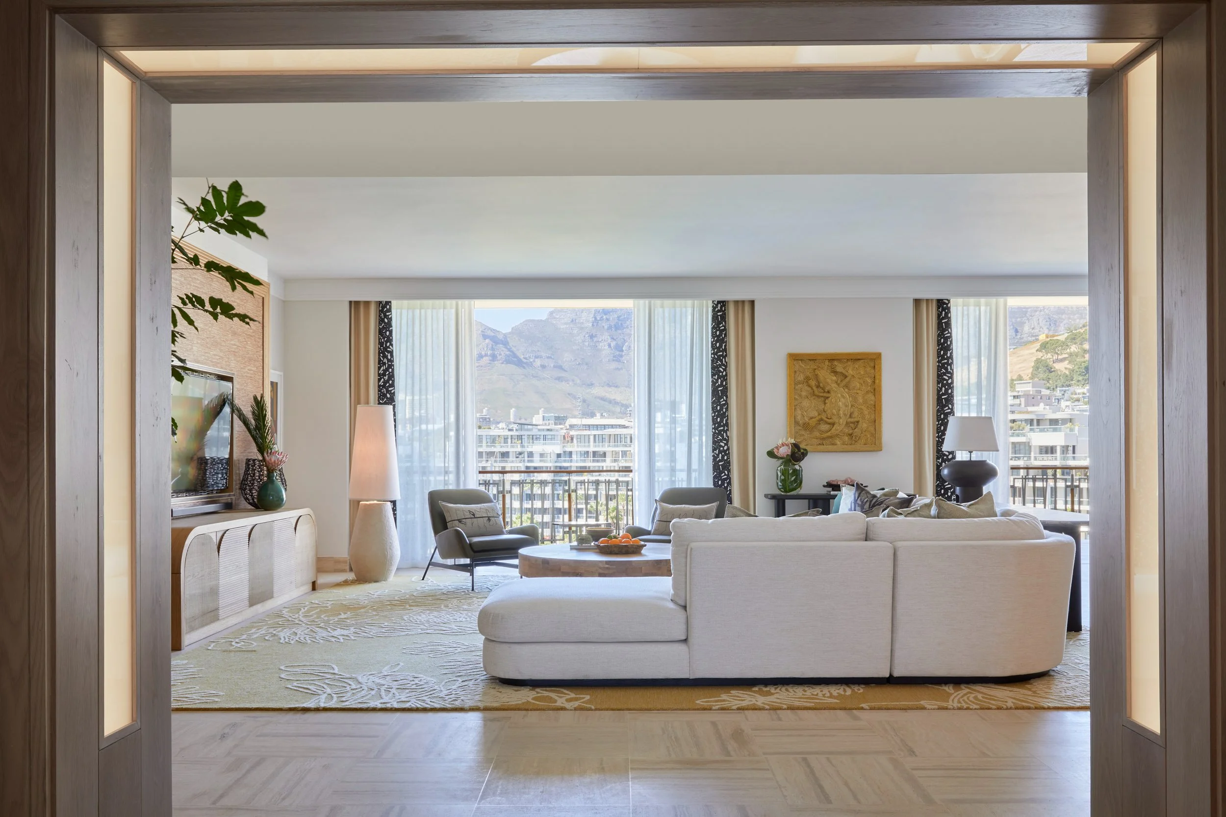 Living room with beige sofas, black chairs, coffee table with fruit, tall floor lamp, TV, plants, wall art, large windows with city view and mountains, decorative curtains, and a patterned rug.