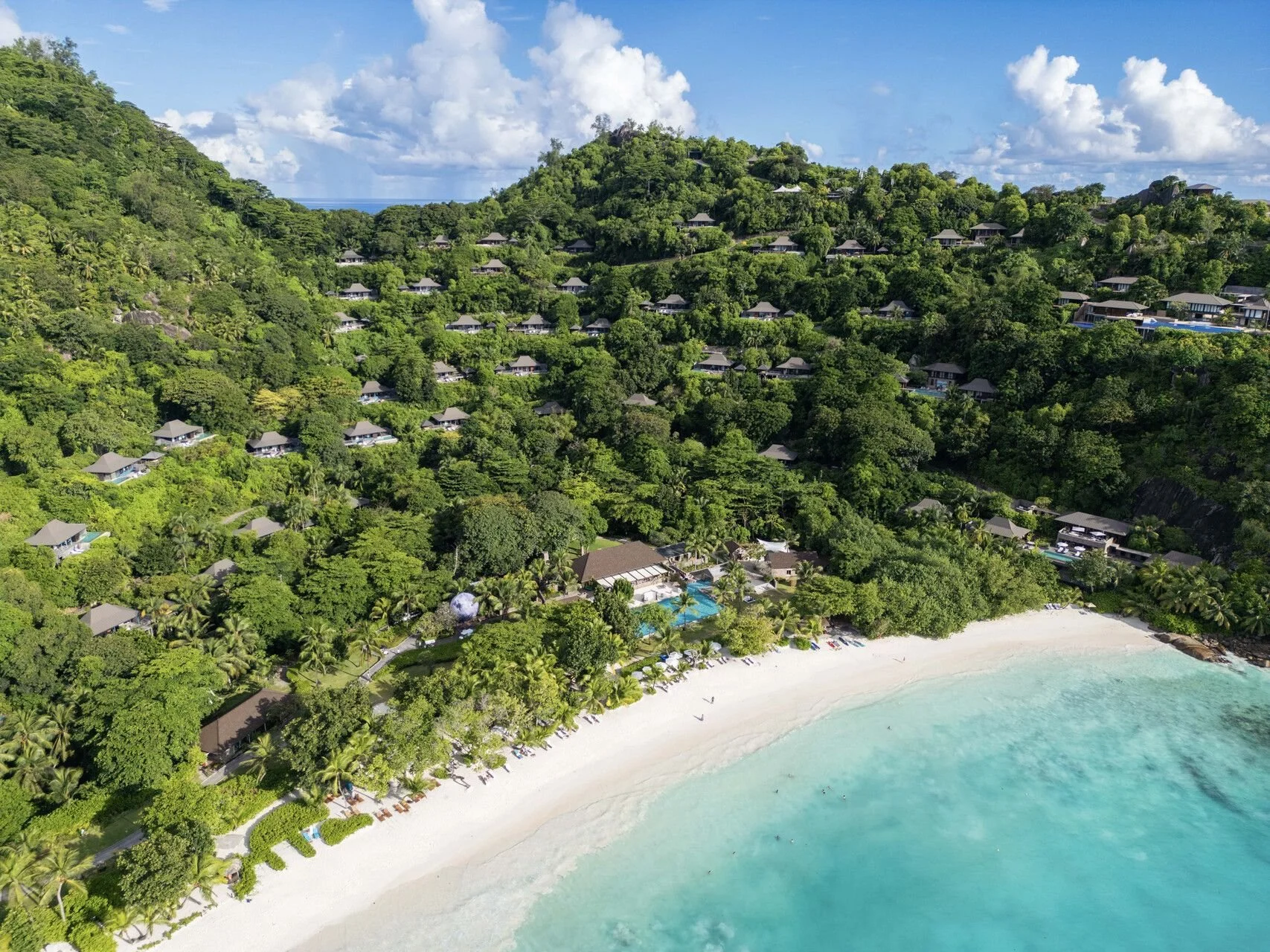 Aerial view of a tropical beach resort with white sandy beach and turquoise water, lined with palm trees and beachfront lounge chairs, adjacent to lush green forested hills with numerous small bavilions or cottages scattered among the trees.