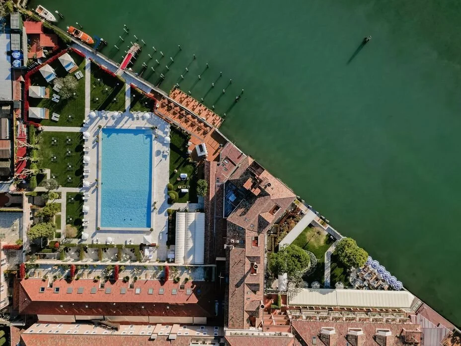 Aerial view of a hotel with a swimming pool, surrounded by buildings, gardens, and docks along a body of water with boats.