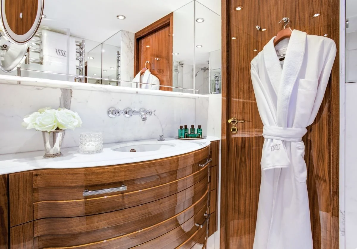 Luxurious bathroom with wooden cabinetry, marble countertops, and a large mirror. A white bathrobe hangs on a hook on a wooden door. Small bottles of toiletries are arranged on the counter, and a vase with white flowers is placed beside the sink.