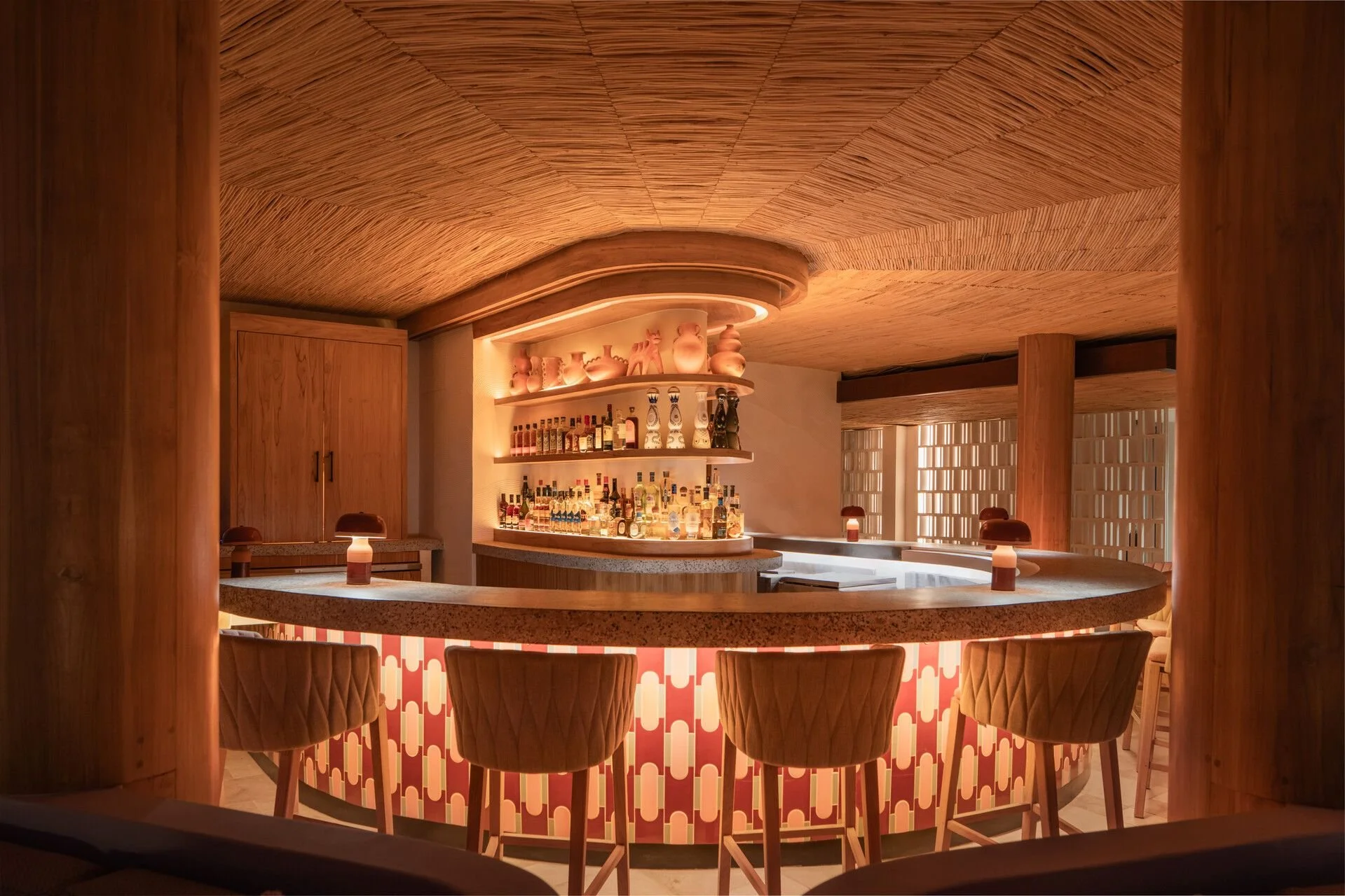 A modern, stylish bar area with a curved marble counter, surrounded by beige quilted bar stools. Shelves behind the bar display various bottles of alcohol and decorative vases, all within a warm, ambient lighting setting with wooden accents.