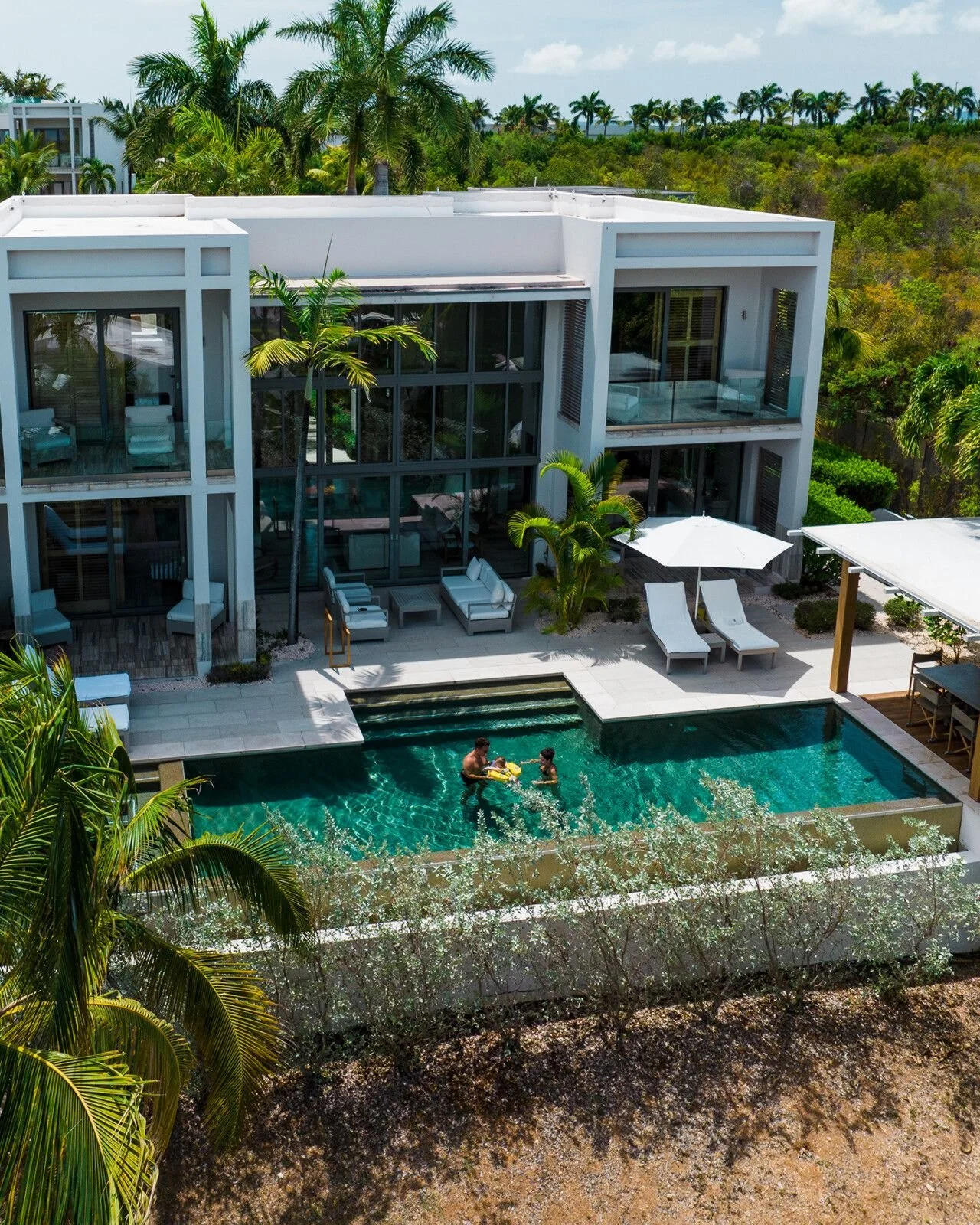 A modern white multi-story house with large glass windows and a pool in the backyard, where two children are playing and adults are supervising. There are lounge chairs with umbrellas, surrounded by lush green palm trees and tropical vegetation.