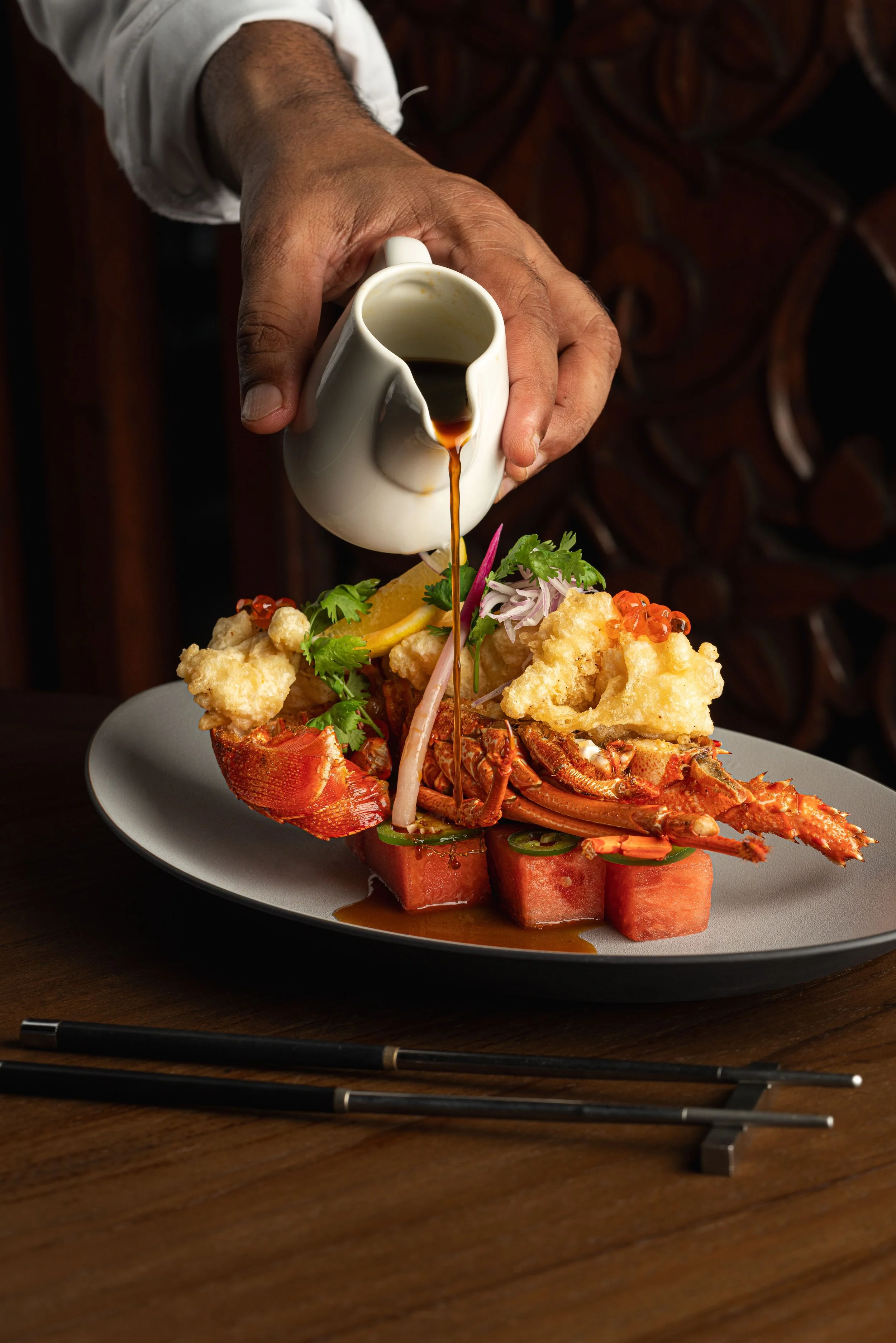 A person pouring soy sauce over a lobster dish garnished with herbs and vegetables on a white plate.