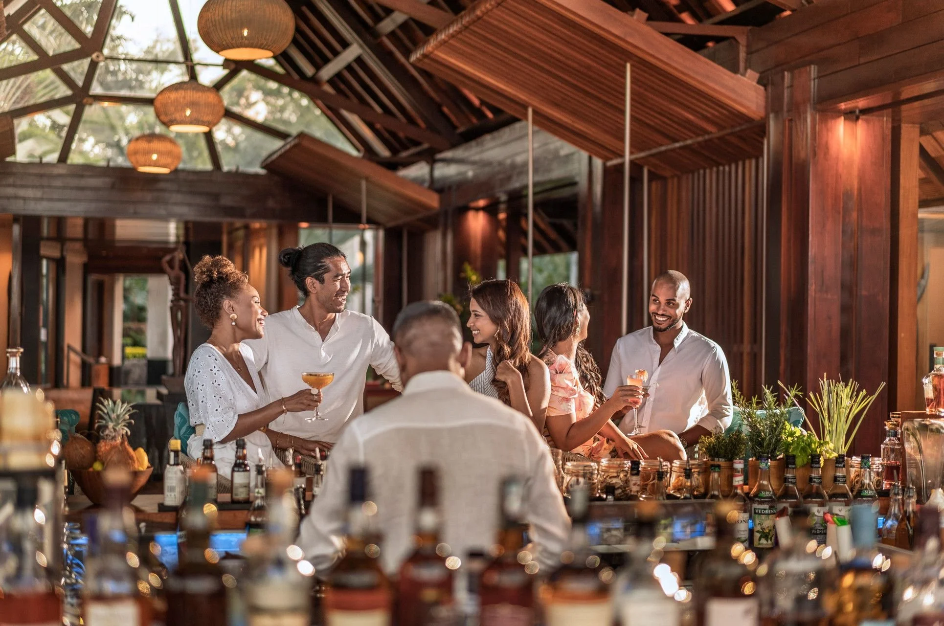 People enjoying a social gathering at a party or celebration in a warm-toned, wooden indoor space with hanging lights. Some individuals are holding drinks and smiling, engaging in conversation.