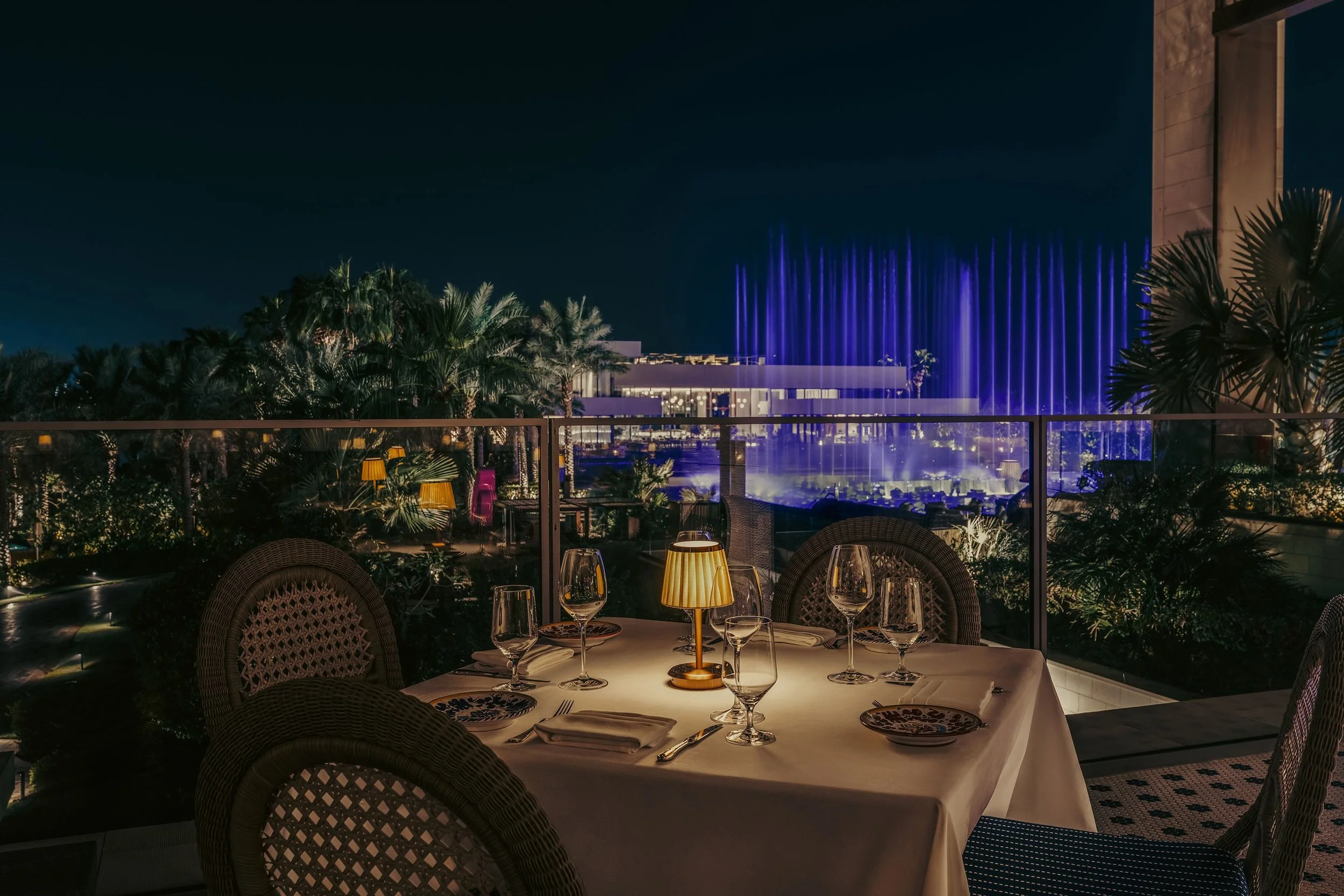 A nighttime outdoor dining patio with a table set for a meal, illuminated by small lamps. In the background, there are lush green trees and a building with blue-lit water fountains. The setting is cozy and elegant.