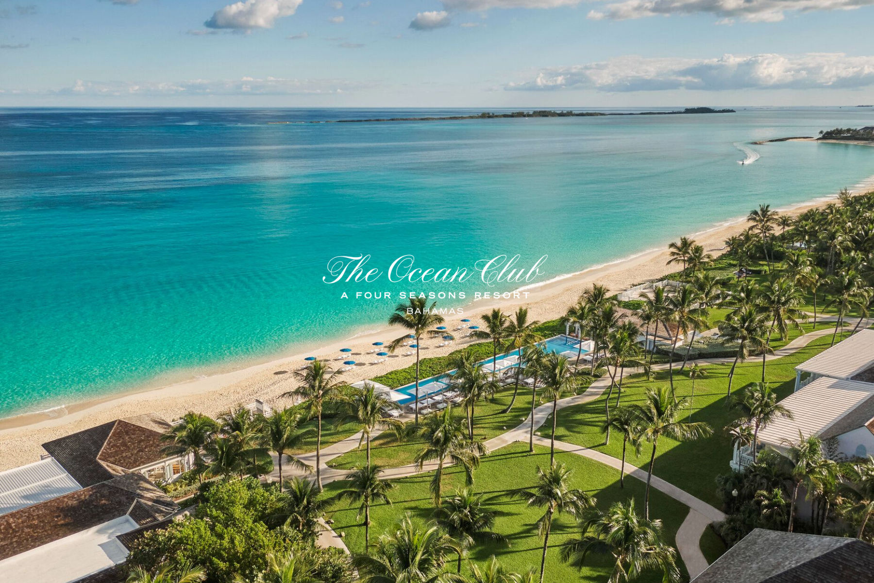 Aerial view of a tropical beach resort with clear turquoise waters, white sandy beach, palm trees, and resort buildings in the foreground. The ocean extends to the horizon with a small island in the distance. Text overlay reads 'The Ocean Club, A Four Seasons Resort, Bahamas'.