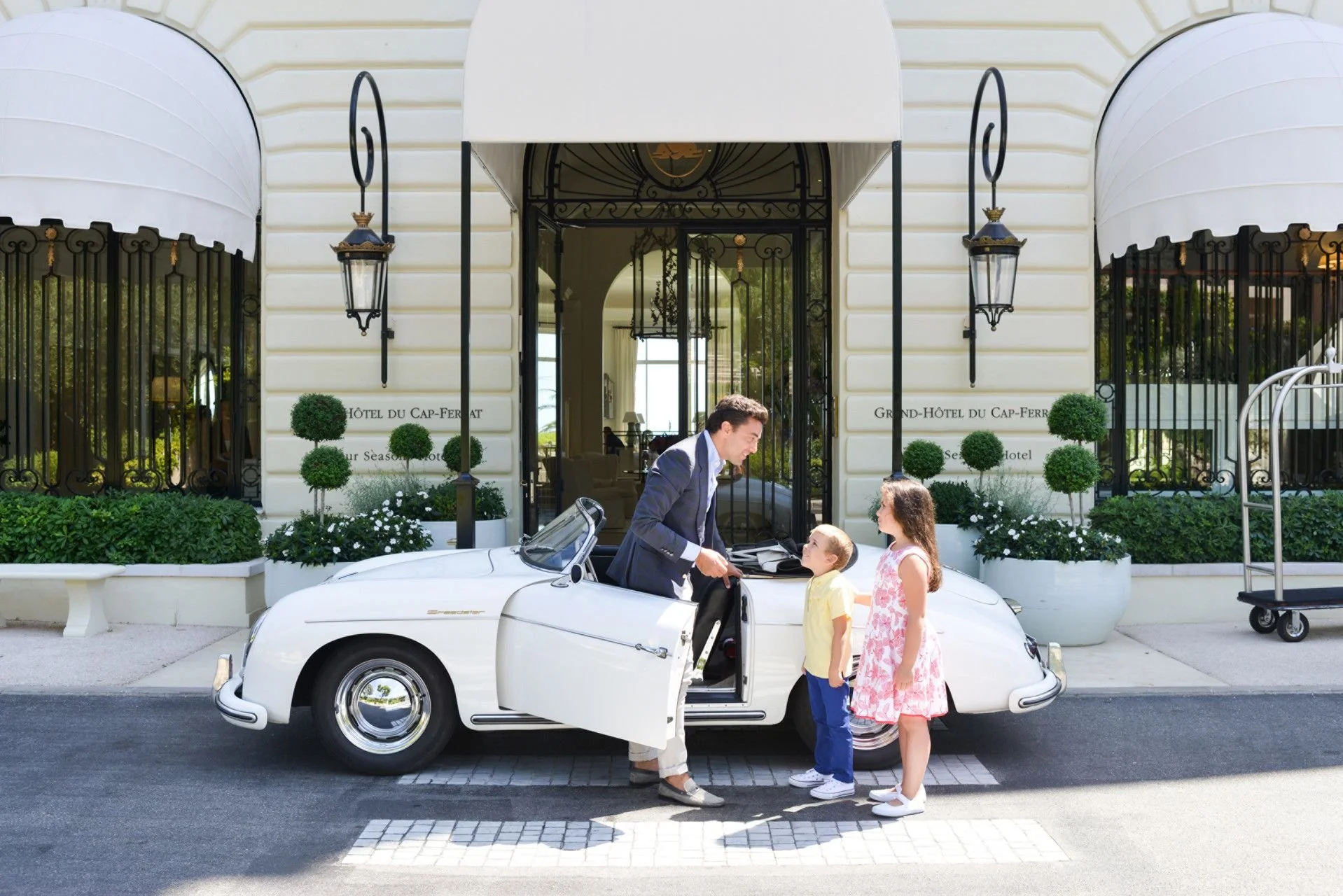 A man in a suit is standing next to a white convertible car, talking to two young children, a boy and a girl, outside a hotel with elegant black iron gates and lanterns. The hotel has a sign reading 'Grand Hôtel du Cap-Ferrat' and 'Pour Saison.'