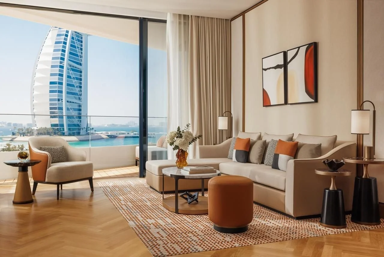 Modern living room with beige sofa, armchair, coffee table, and abstract artwork, overlooking a cityscape with a sail-shaped building.