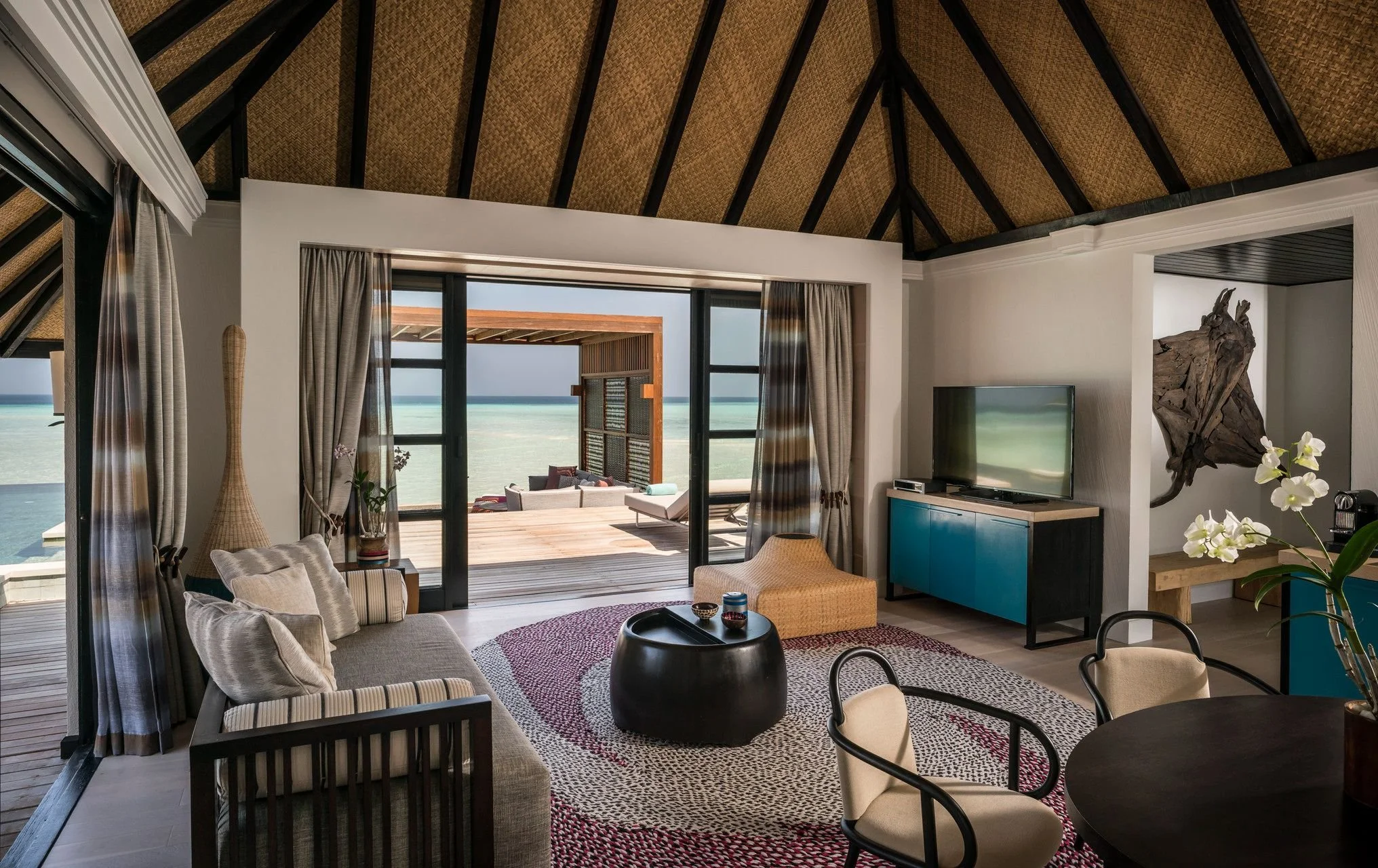 Living room with large sliding glass doors leading to a balcony with ocean view, furnished with chairs, a sofa, a blue cabinet, a flat-screen TV, and decorative items.