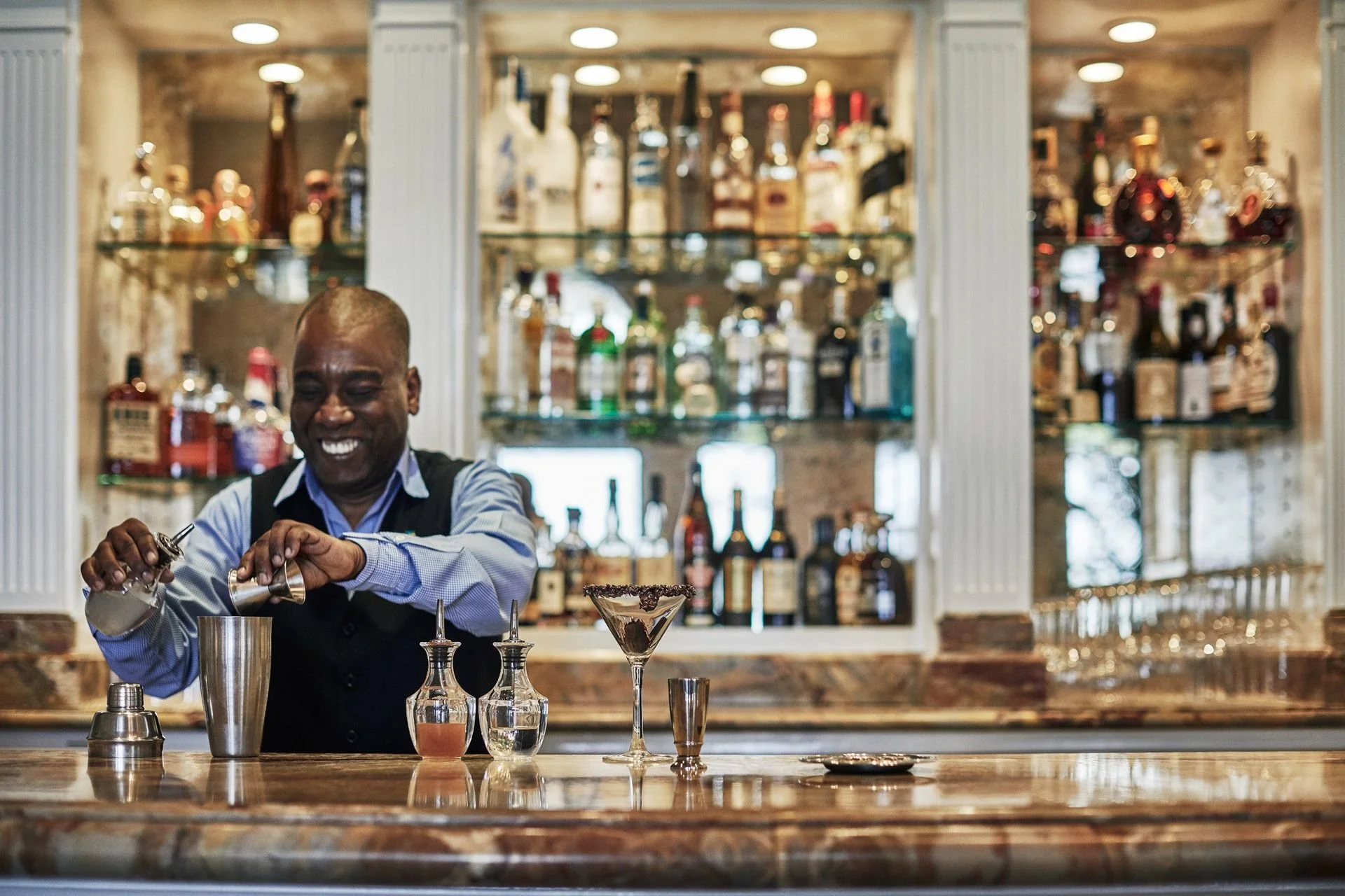 A smiling bartender preparing a cocktail behind a bar, with various bottles of alcohol displayed on shelves in the background.