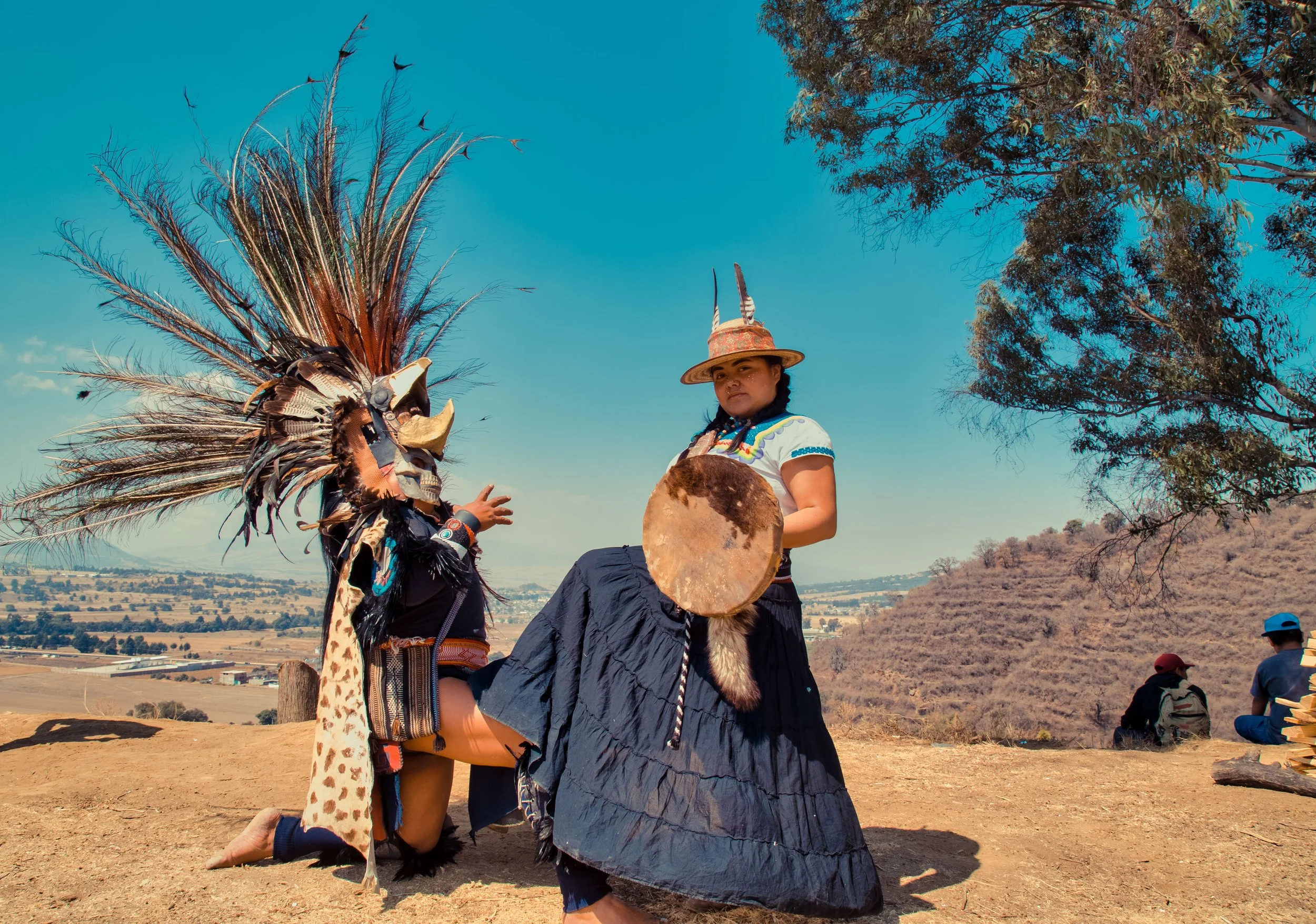Indigenous girl wearing traditional clothing and a feathered headdress, holding a drum, standing outdoors in a rural landscape under a blue sky.