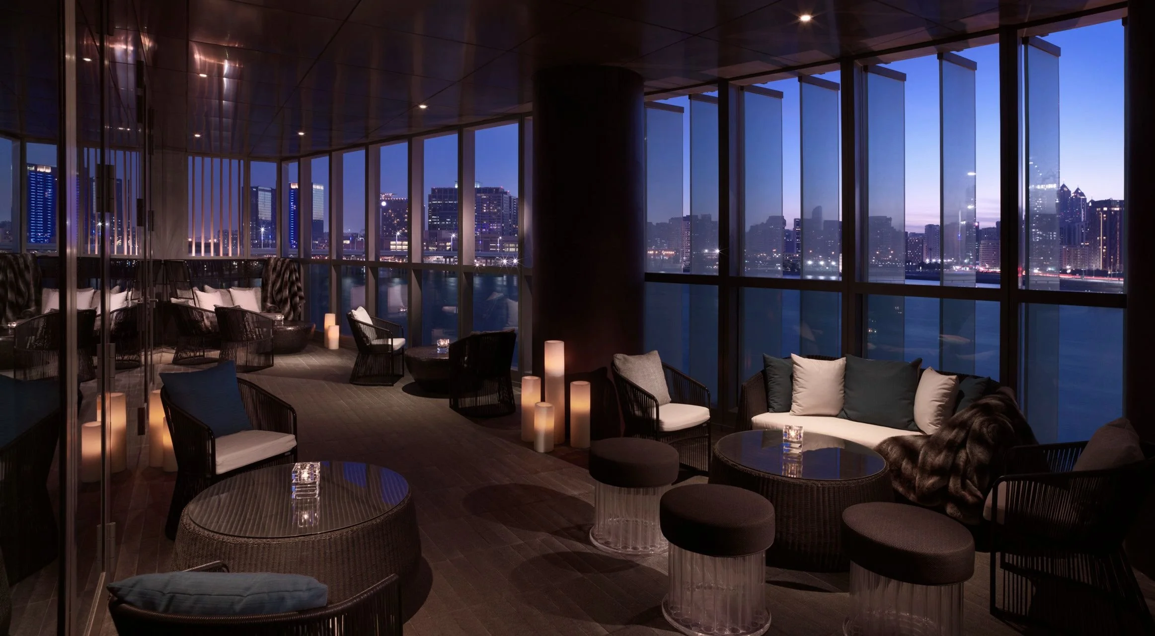 Interior of a modern lounge area with large windows showing a city skyline at dusk, illuminated buildings, and water. The space features contemporary furniture, pillows, candles, and soft lighting.