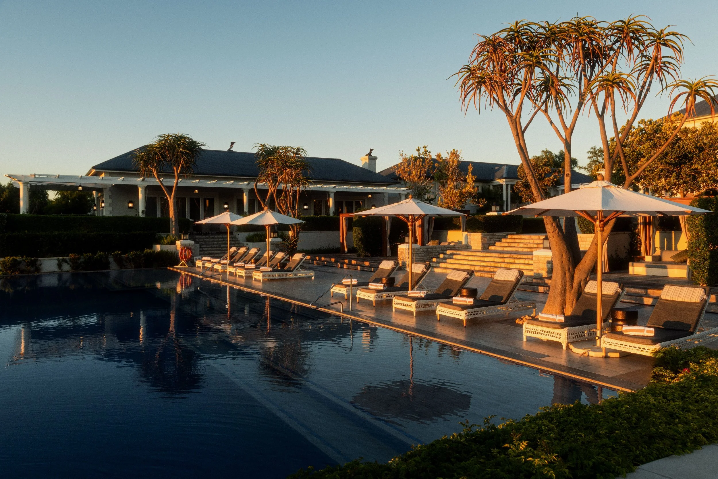 Luxury house with a pool and lounge chairs at sunset, surrounded by trees.