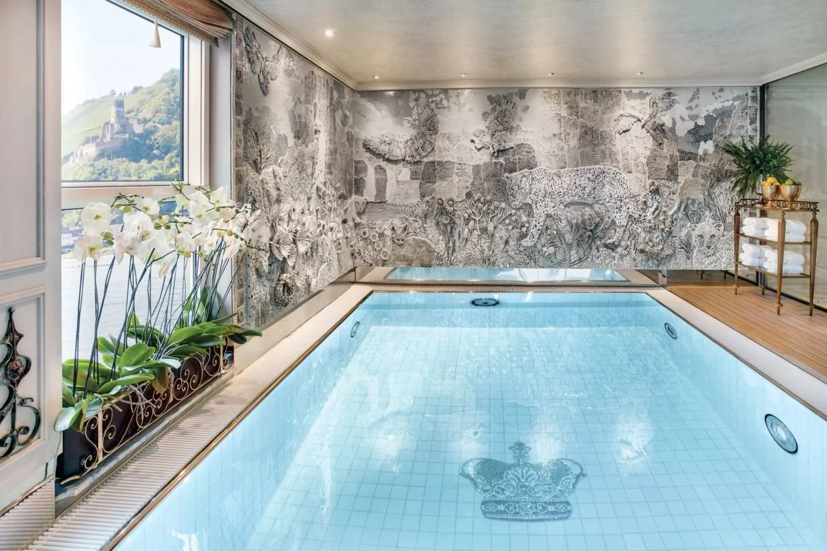 Indoor swimming pool with a mural of animals on the wall, a window with a mountain view, plants, and a small shelving unit with towels.