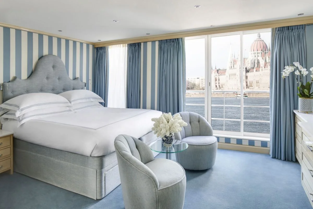 A hotel room with a bed, two chairs around a glass table, and large windows offering a view of a historic building and a river.