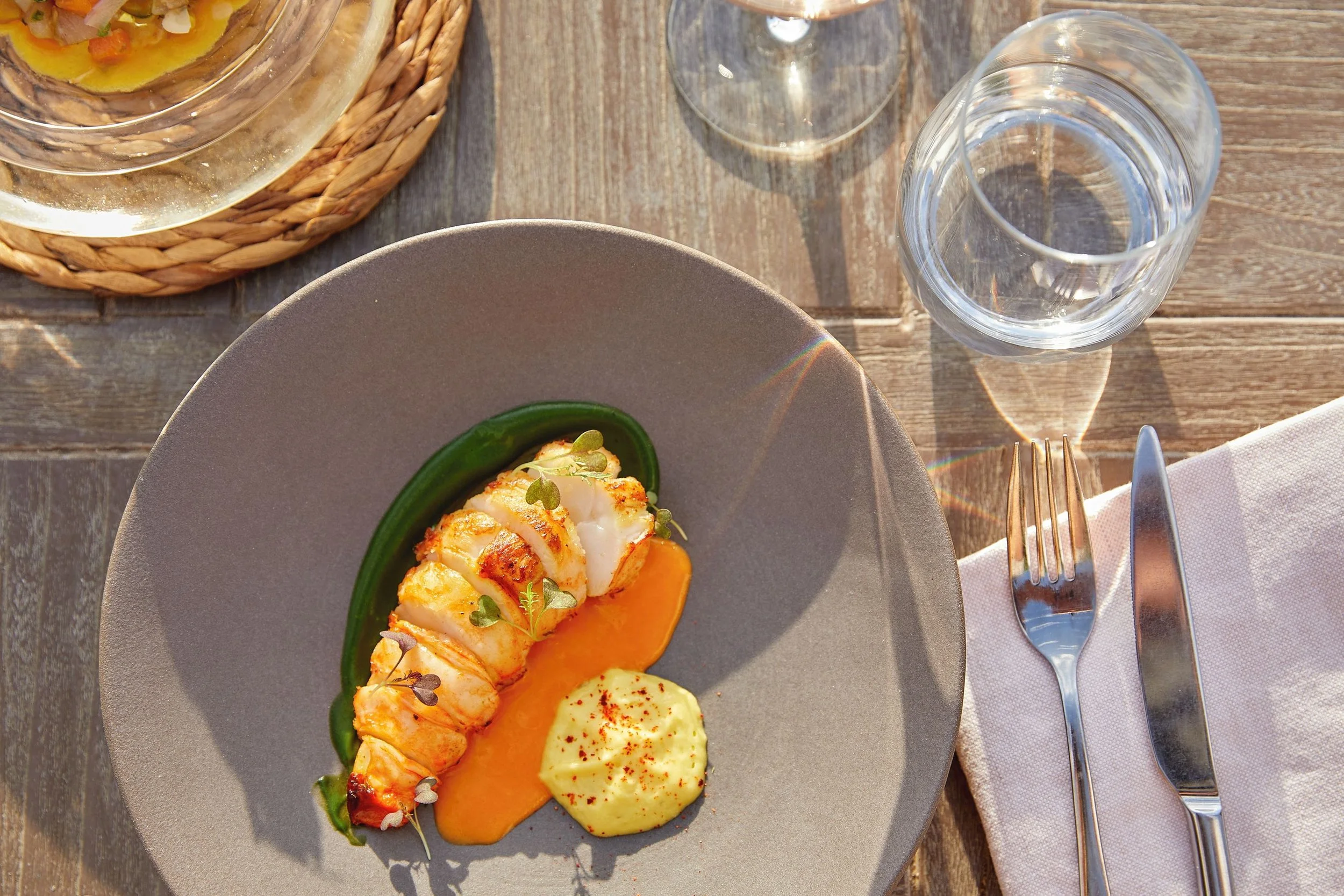 A plate of cooked lobster tail with mashed potatoes, served with a dipping sauce on the side, placed on a wooden table with a glass of water and a wine glass nearby.