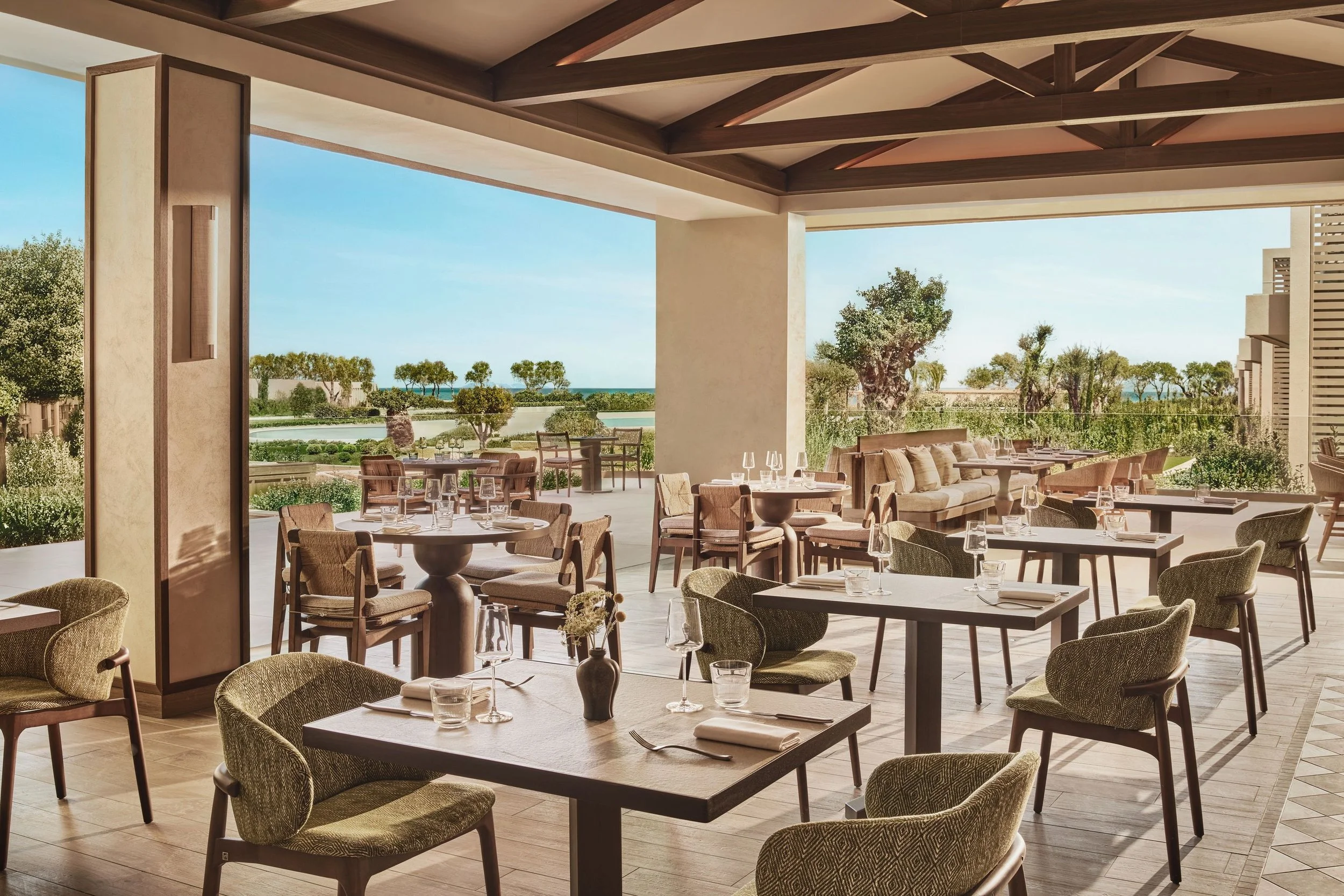 Indoor restaurant or lounge area with tables set with wine glasses and napkins, large open windows revealing trees and a body of water outside, and a wooden ceiling with exposed beams.