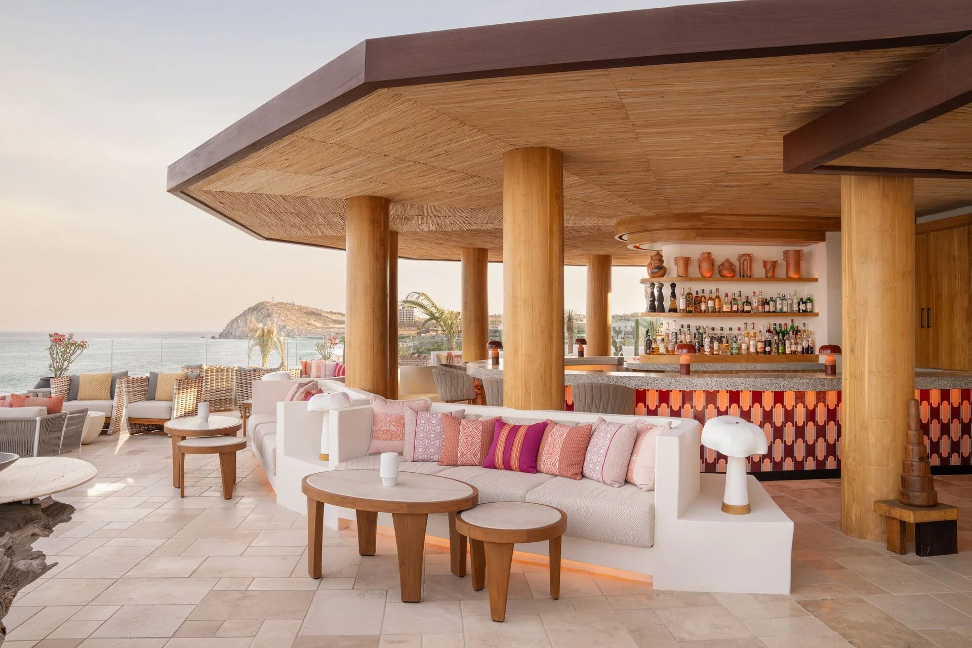Luxury outdoor bar and lounge area with seating, overlooking a beach and ocean with a hill in the background.