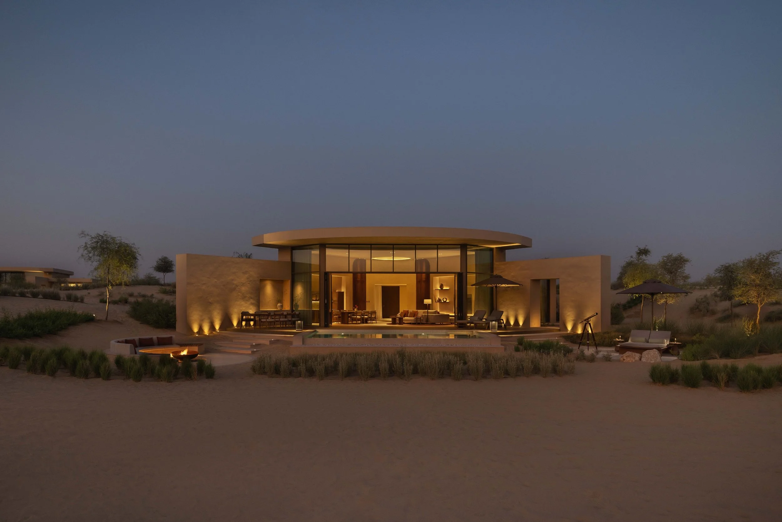 Luxury modern house with large glass windows and indoor lighting, surrounded by desert landscape with sparse trees, during dusk.