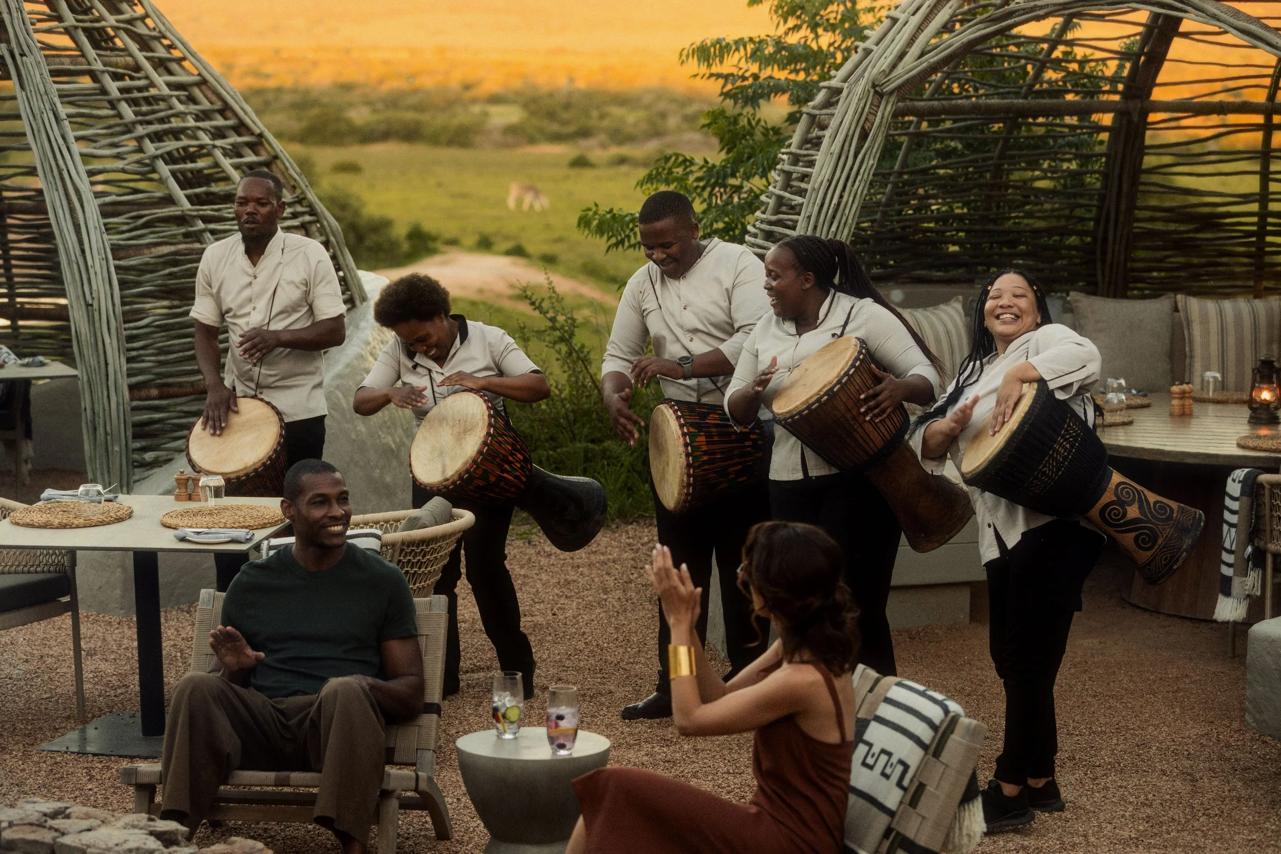 Group of people playing drums and singing outdoors during sunset, with tables and chairs around, and a woven structure in the background.