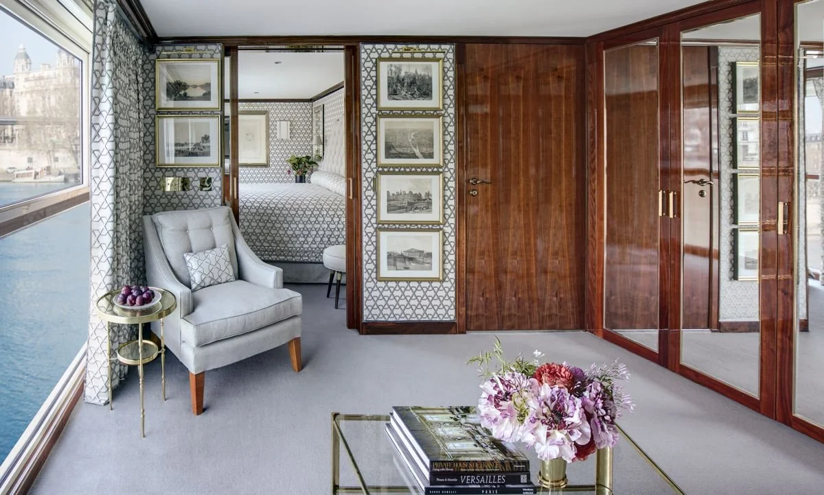 A luxurious hotel room with large windows overlooking a river, a comfortable gray armchair with a small pillow, a side table with grapes, a glass coffee table with a vase of flowers and books, patterned wallpaper, wood-paneled sliding doors with mirrored panels, and framed black-and-white artwork.