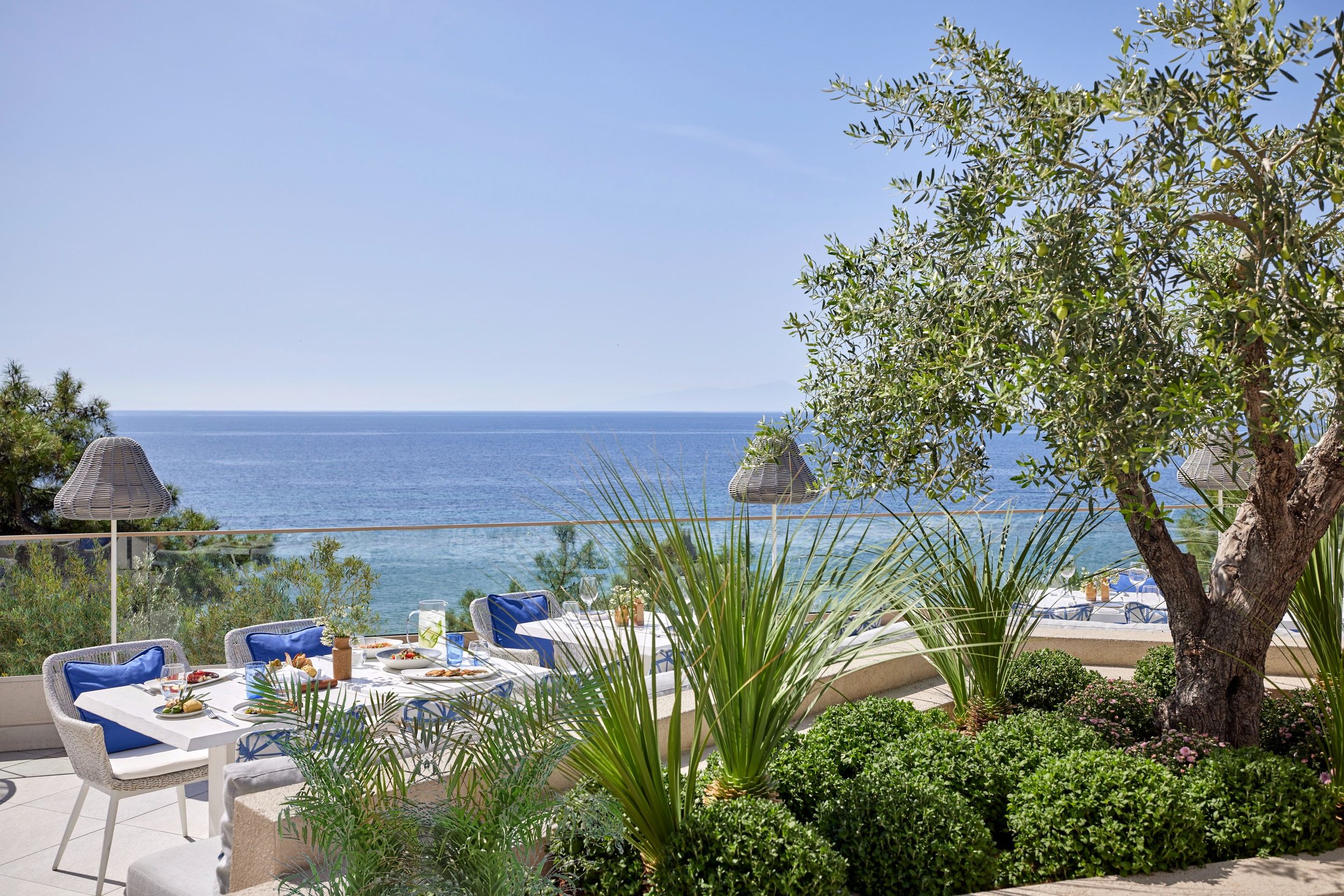 Outdoor restaurant setup with a view of the ocean, surrounded by lush green plants and trees, with tables and chairs set for dining.