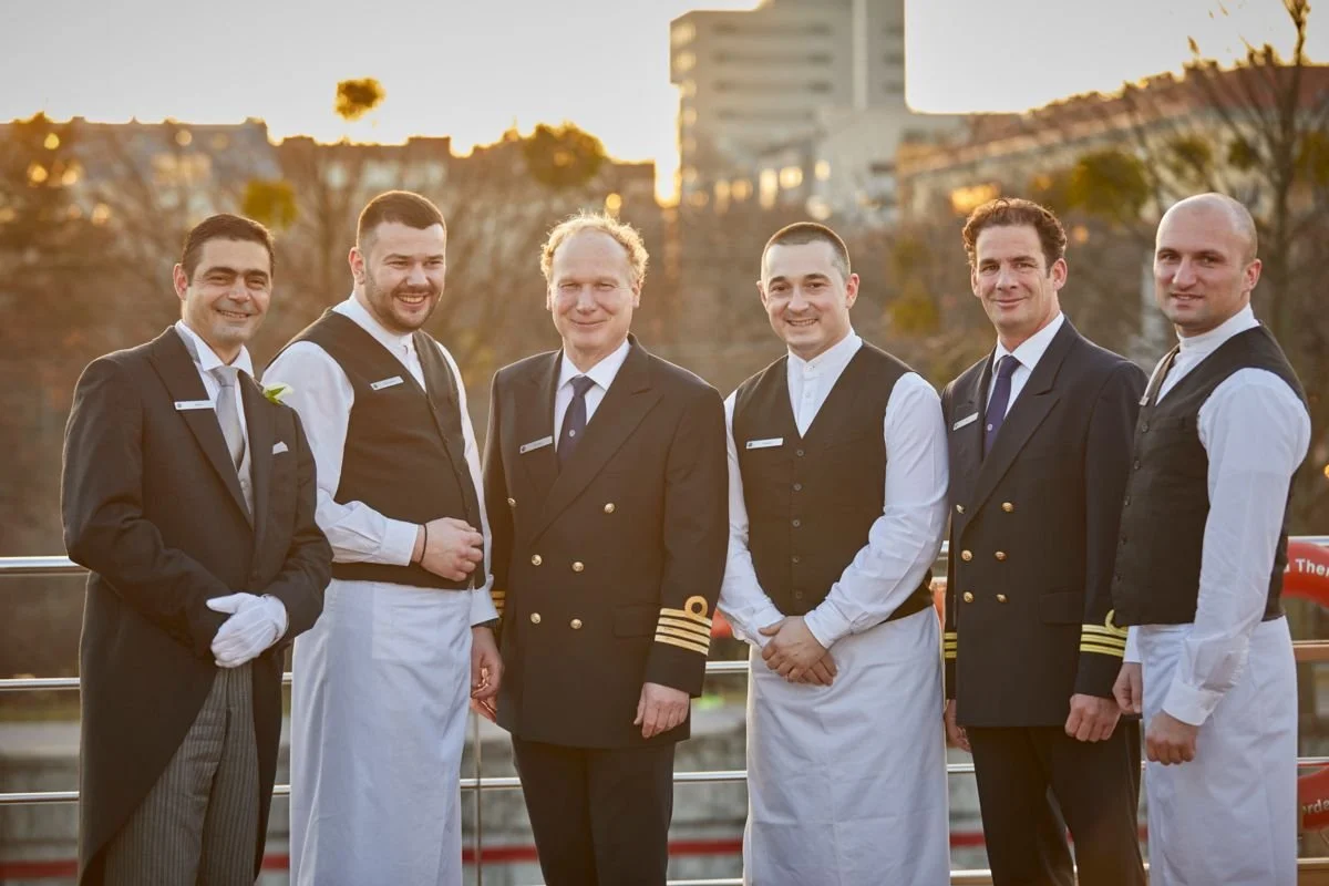 Seven men dressed in formal uniforms, including some with suits and others with chef uniforms, standing outdoors during sunset, smiling at the camera.