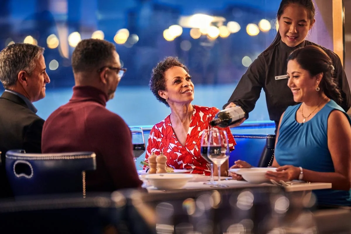 A woman in a red and white floral dress smiling as she gets served wine at a restaurant table with three other people, two women and one man, and a waitress pouring wine during the evening.
