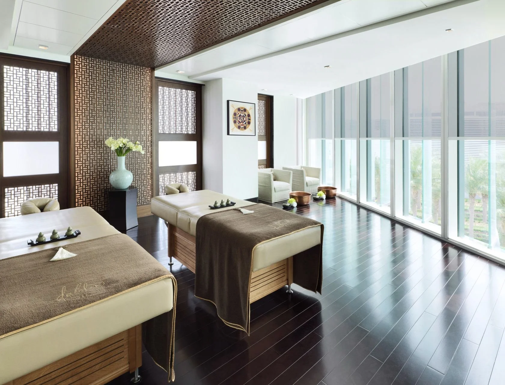 Spa room with massage tables, dark wooden flooring, large windows with blinds, and modern decor including framed artwork and a tall vase with white flowers.