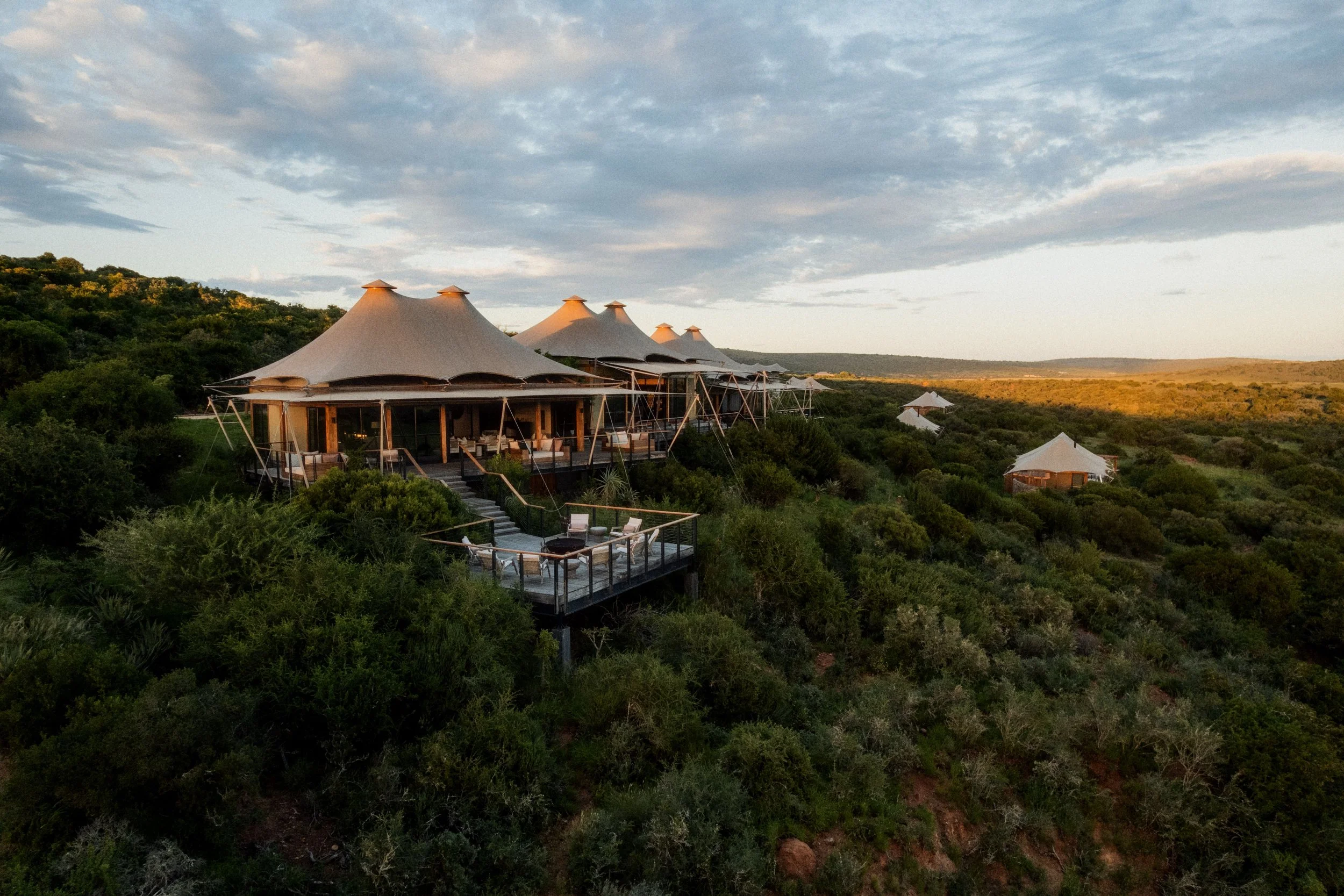 Luxury tents with pointed roofs on a hillside surrounded by greenery under a partly cloudy sky