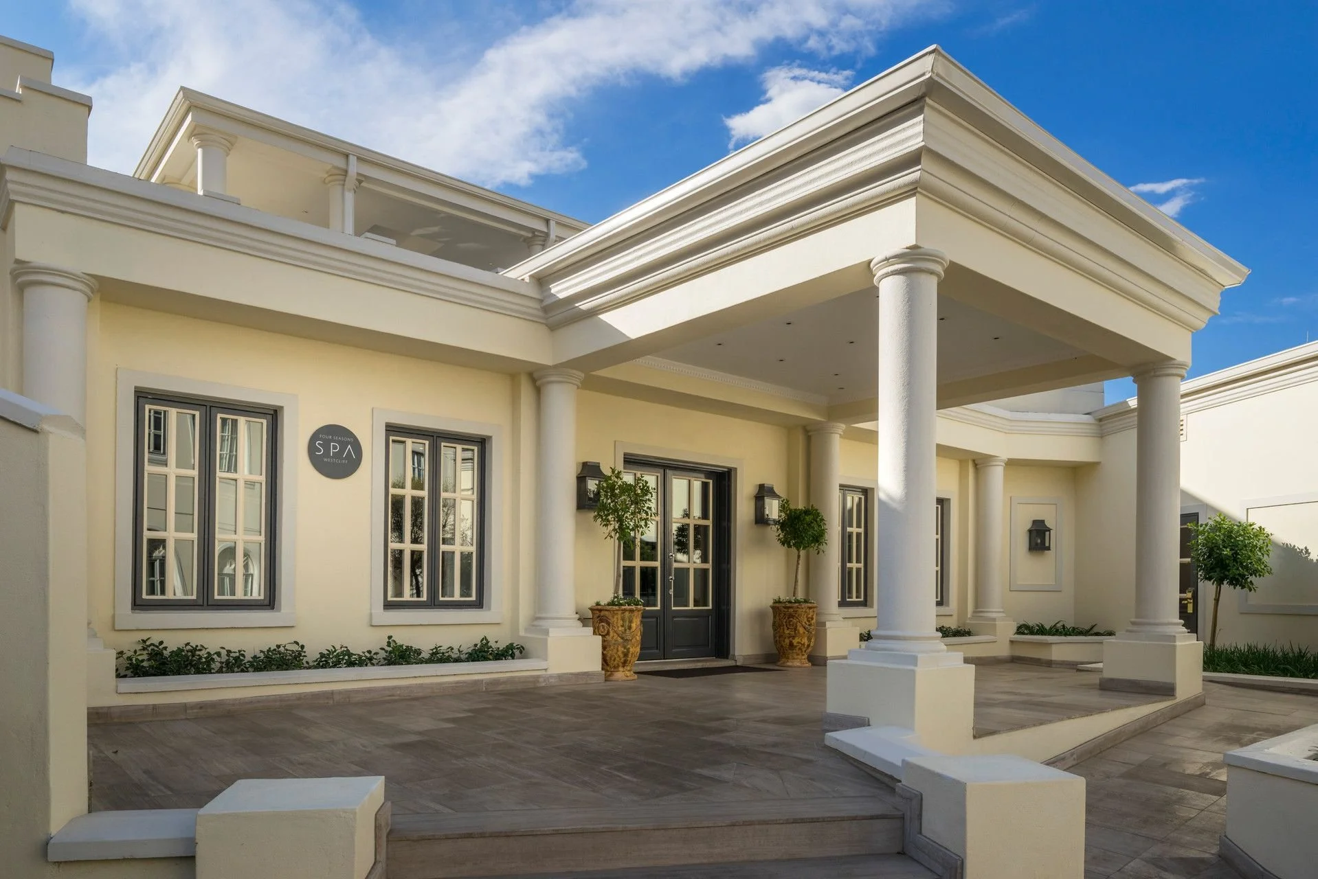 Front view of a luxury building with cream-colored walls, white columns, and black-framed windows. Potted plants and wall-mounted lanterns decorate the entrance under a clear blue sky.