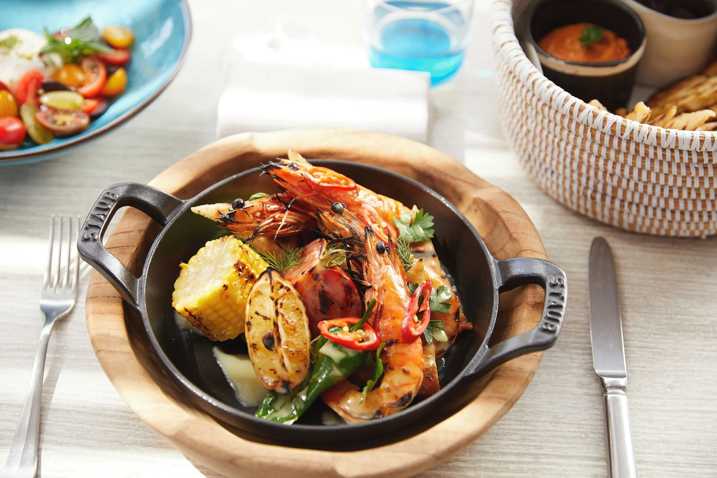 Seafood dish with shrimp, pineapple, and greens served in a small black skillet on a wooden plate, with side dishes including salad and dips on a table.