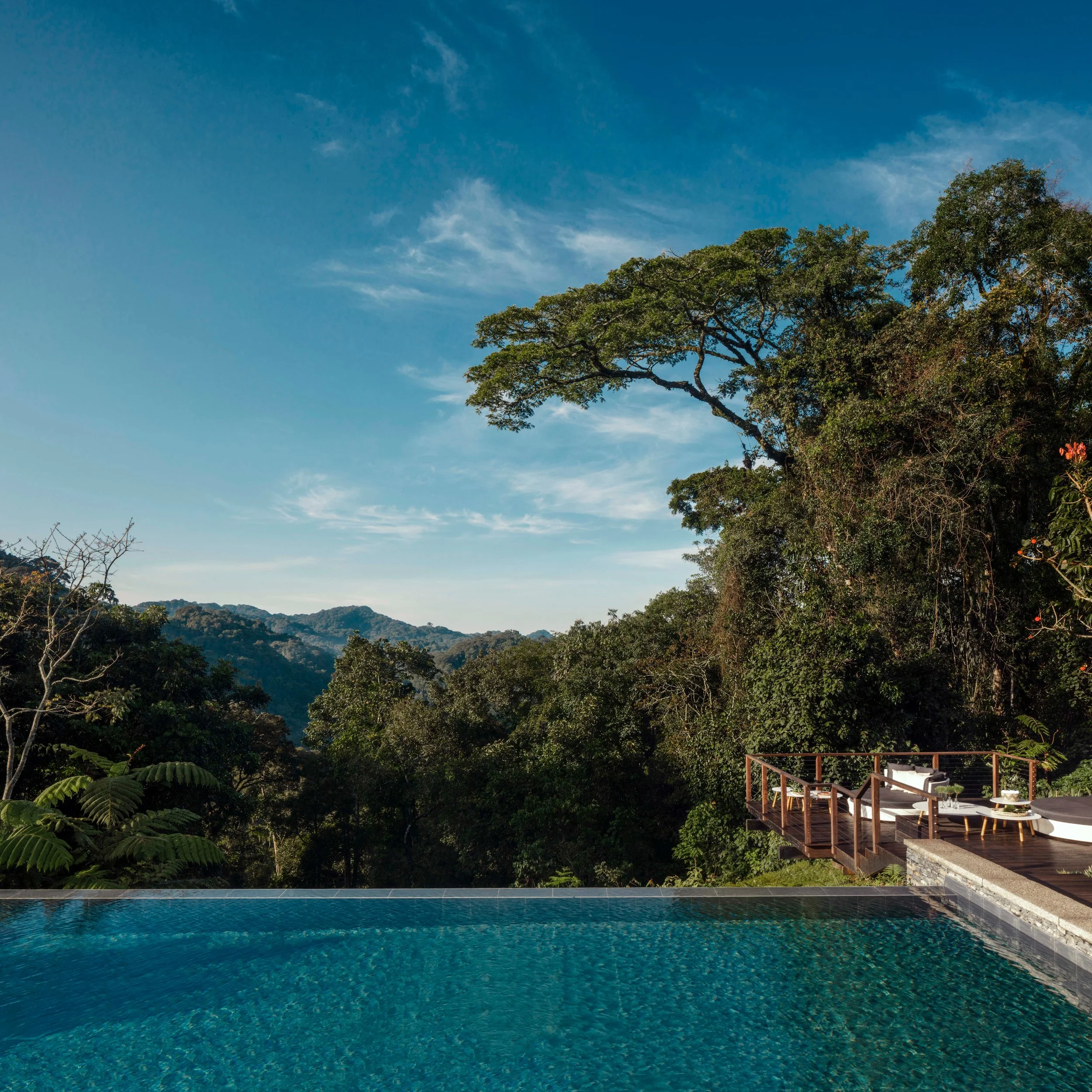 View of an outdoor swimming pool with a scenic backdrop of green mountains and trees, and a hillside terrace with seating area.