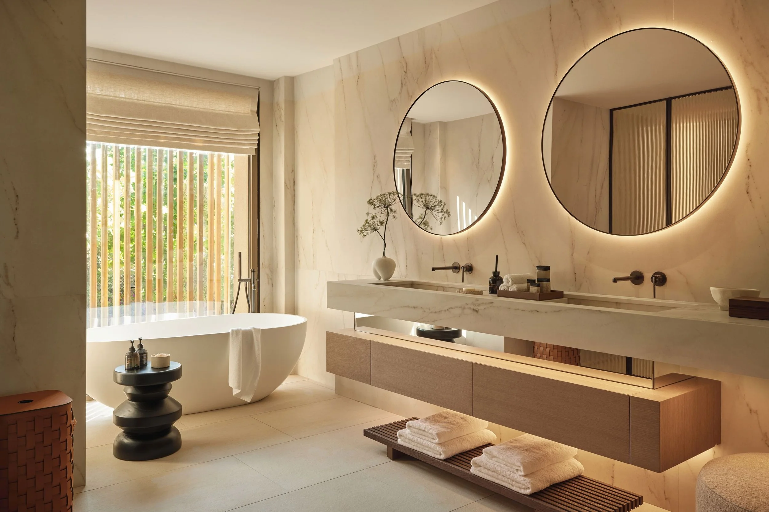 Modern bathroom featuring a freestanding bathtub near a window with beige blinds, two illuminated round mirrors above a marble vanity with folded towels on a shelf, decorative plant, soap dispensers, and a stool.