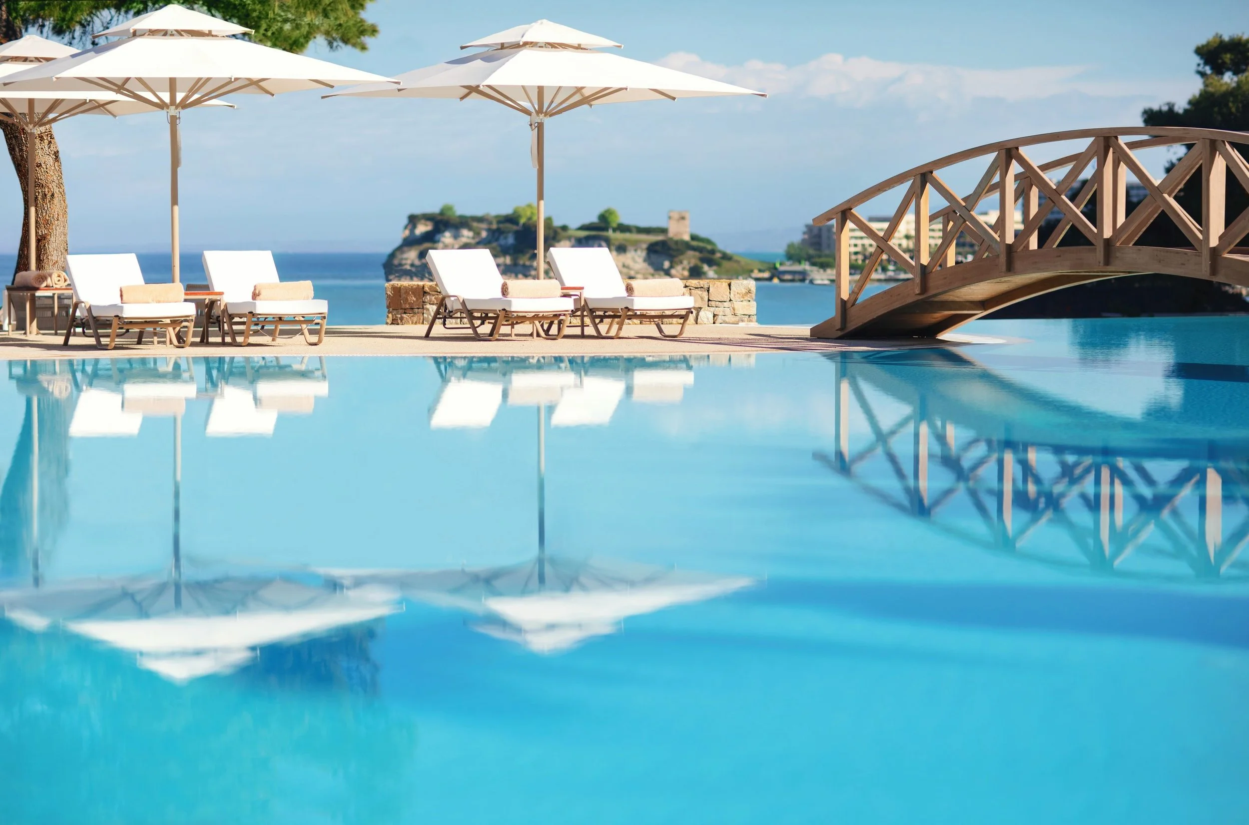 A serene outdoor pool area with white lounge chairs, large white umbrellas, and a small wooden bridge over the pool, overlooking a view of cliffs, water, and a partly cloudy sky.