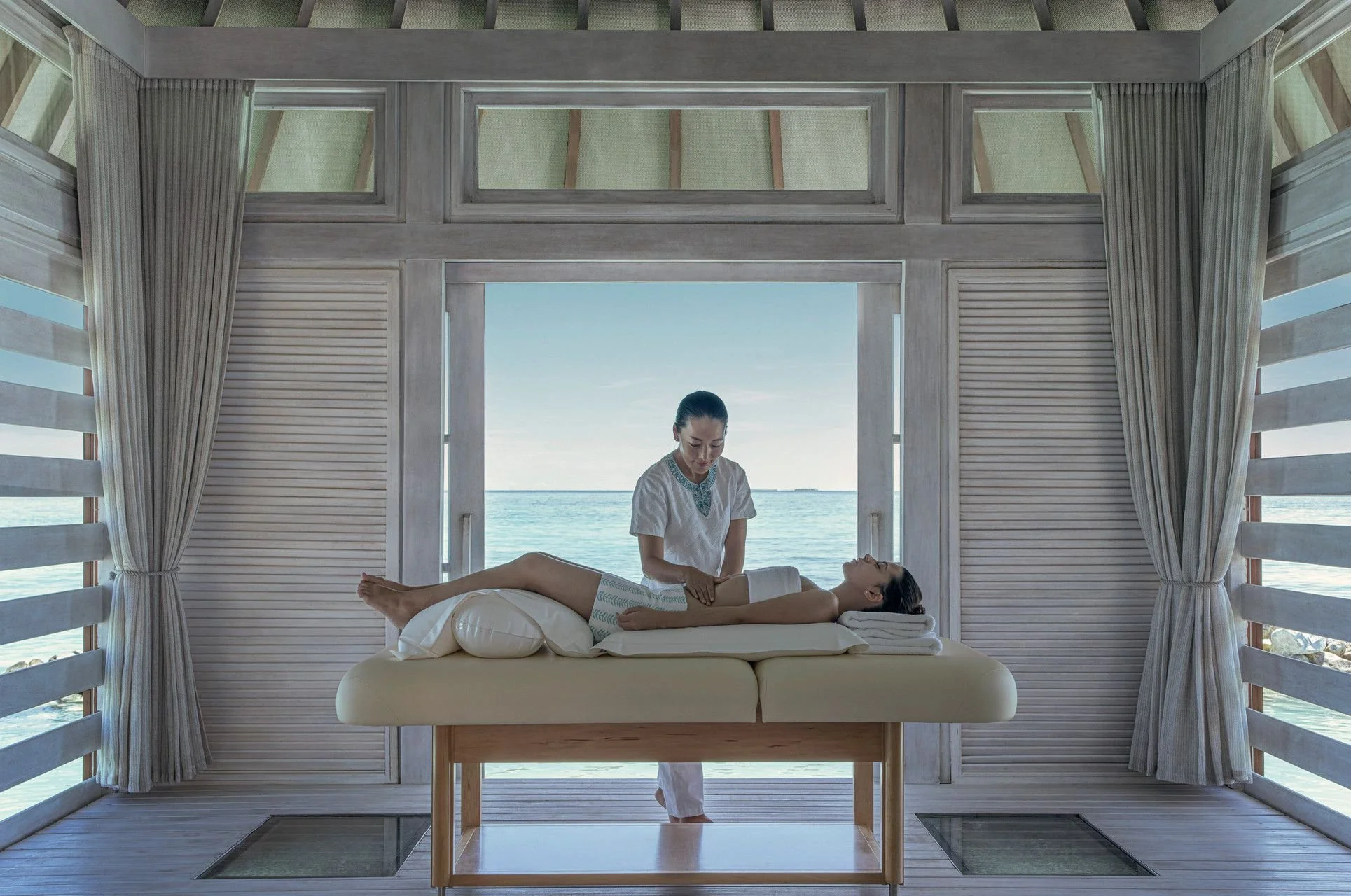 A woman is receiving a massage on a massage table by a therapist at a seaside spa with large windows showing the ocean.